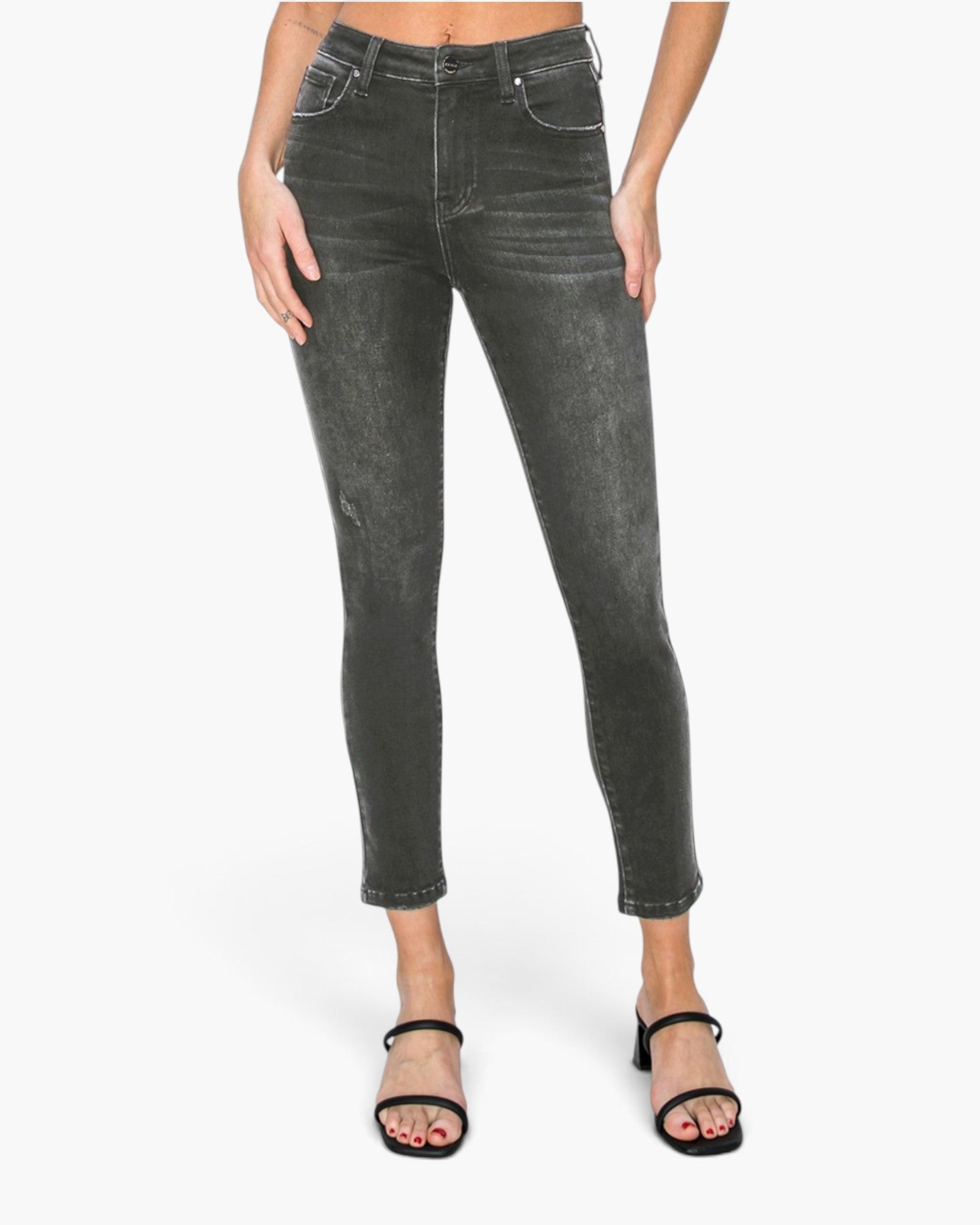A model wearing dark gray washed skinny jeans with a cropped ankle length, paired with black strappy sandals, shown against a plain white studio background.