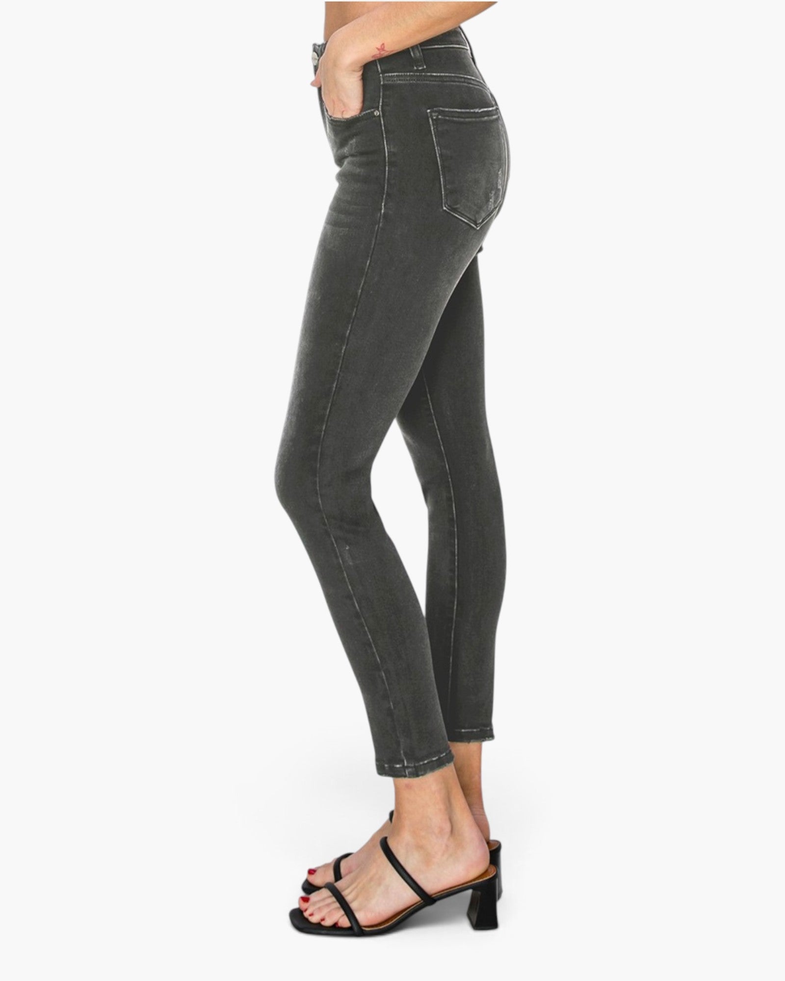 A side profile view of a person wearing dark charcoal gray skinny jeans and black strappy heeled sandals against a plain white background.