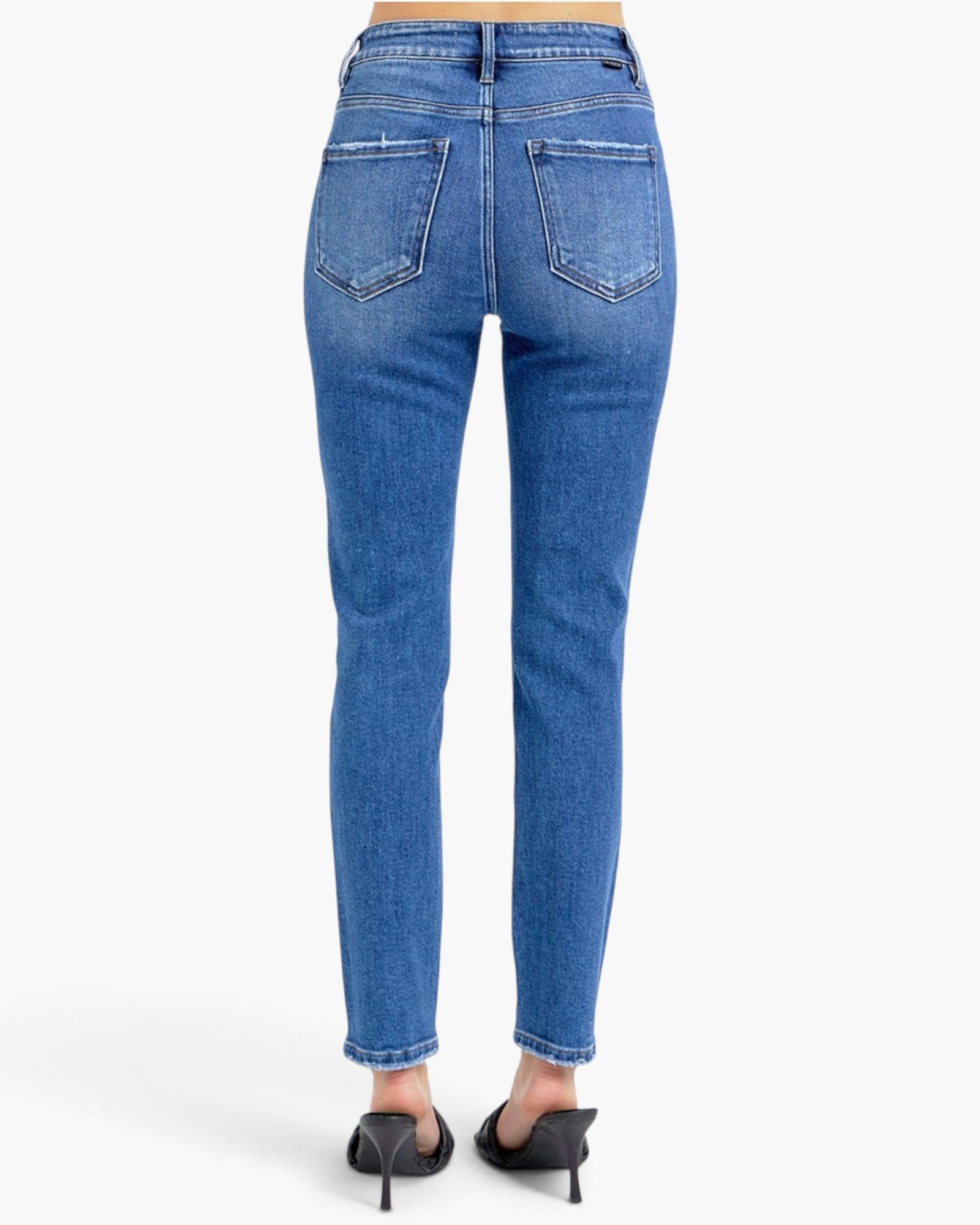 Rear view of a person wearing medium-wash blue skinny jeans with black pointed-toe heels against a plain white studio background.