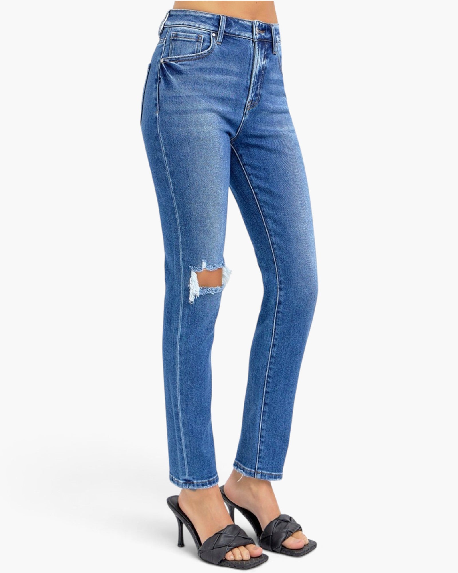 A person wearing blue skinny jeans with a distressed rip on the left thigh, paired with black heeled slide sandals, shown against a plain light gray studio background.