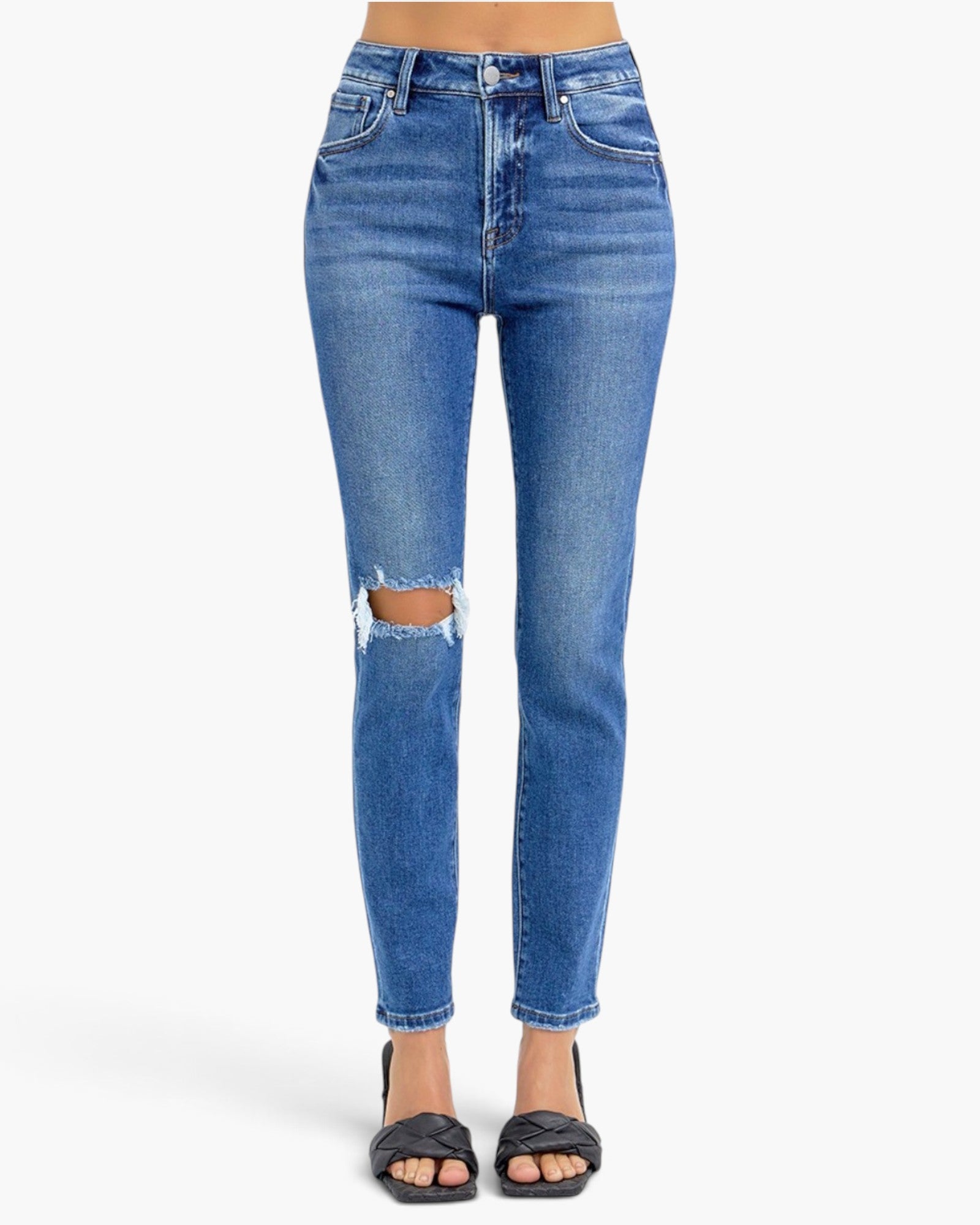 A model's lower body wearing medium wash blue denim jeans with a high-rise fit, ripped knees, and a cropped ankle length, paired with black slide sandals against a plain white background.