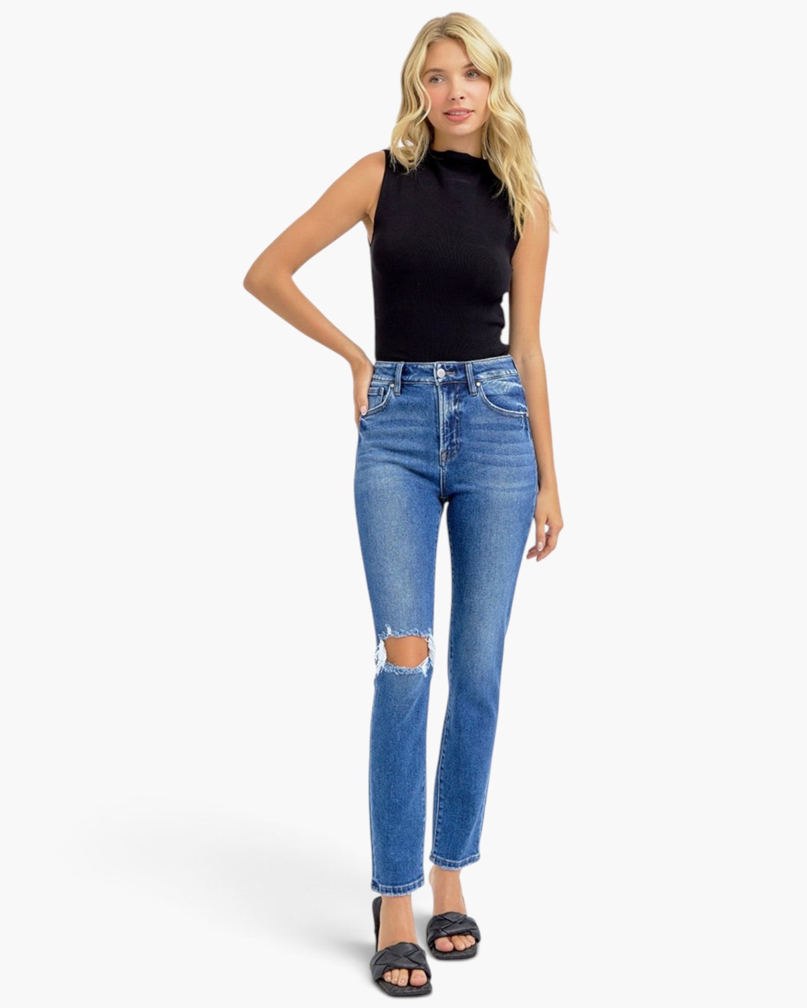 A blonde model wearing a black sleeveless mock-neck top, blue high-waisted skinny jeans with a distressed knee, and black slide sandals, standing against a light gray studio background.