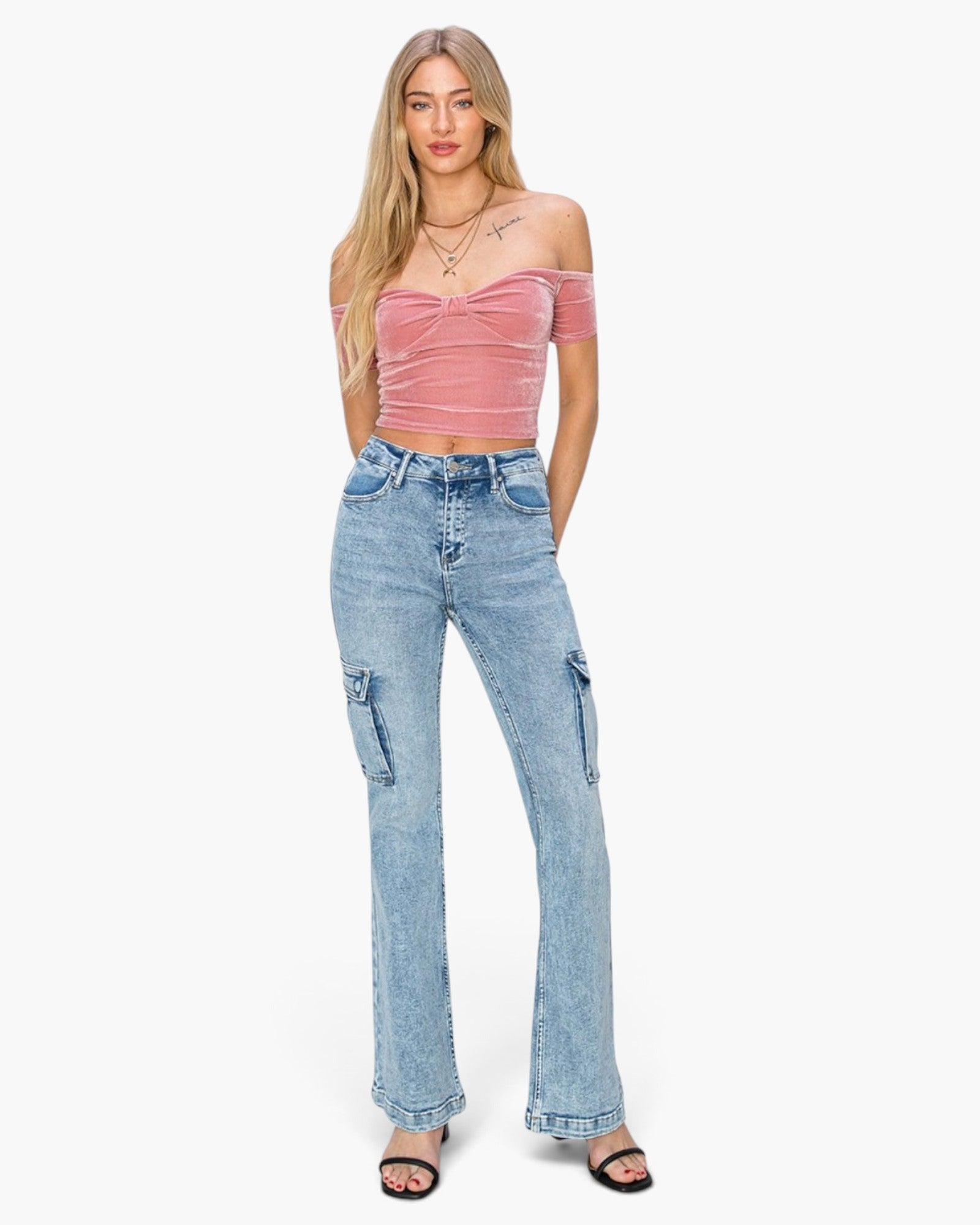 A woman modeling a dusty pink off-the-shoulder velvet crop top and light-wash blue high-rise flare jeans with cargo pockets, paired with black slide sandals, against a plain white background.
