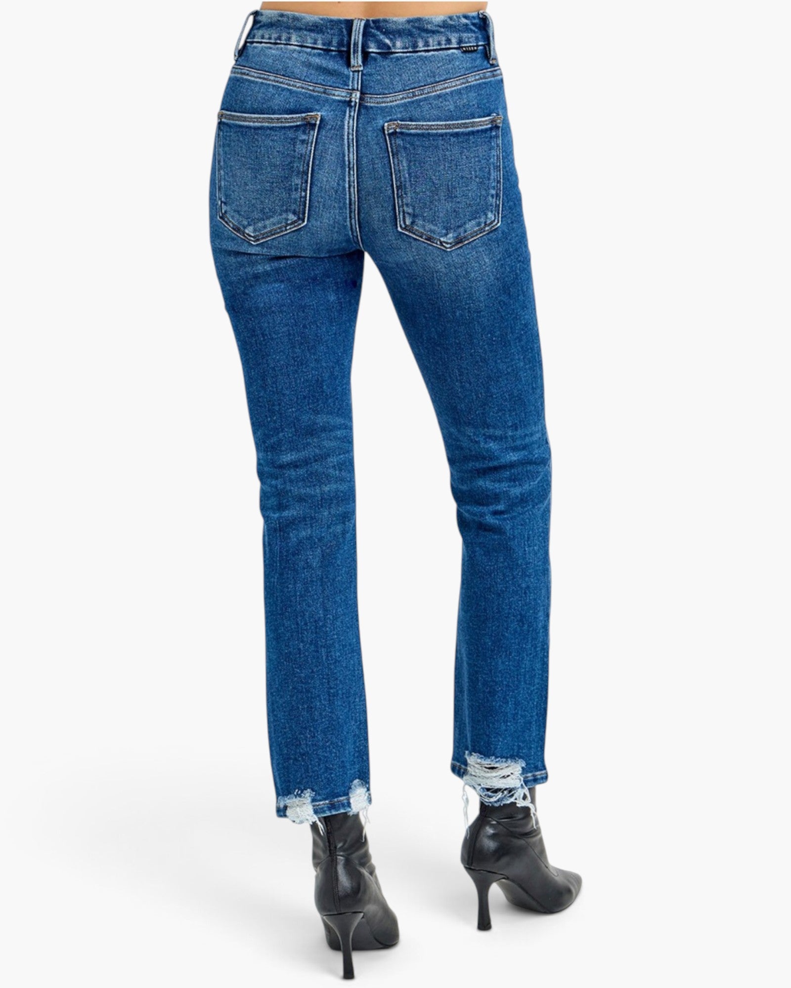 Rear view of a person wearing blue denim jeans with a raw, frayed hem and black heeled ankle boots, shown against a plain white studio background.