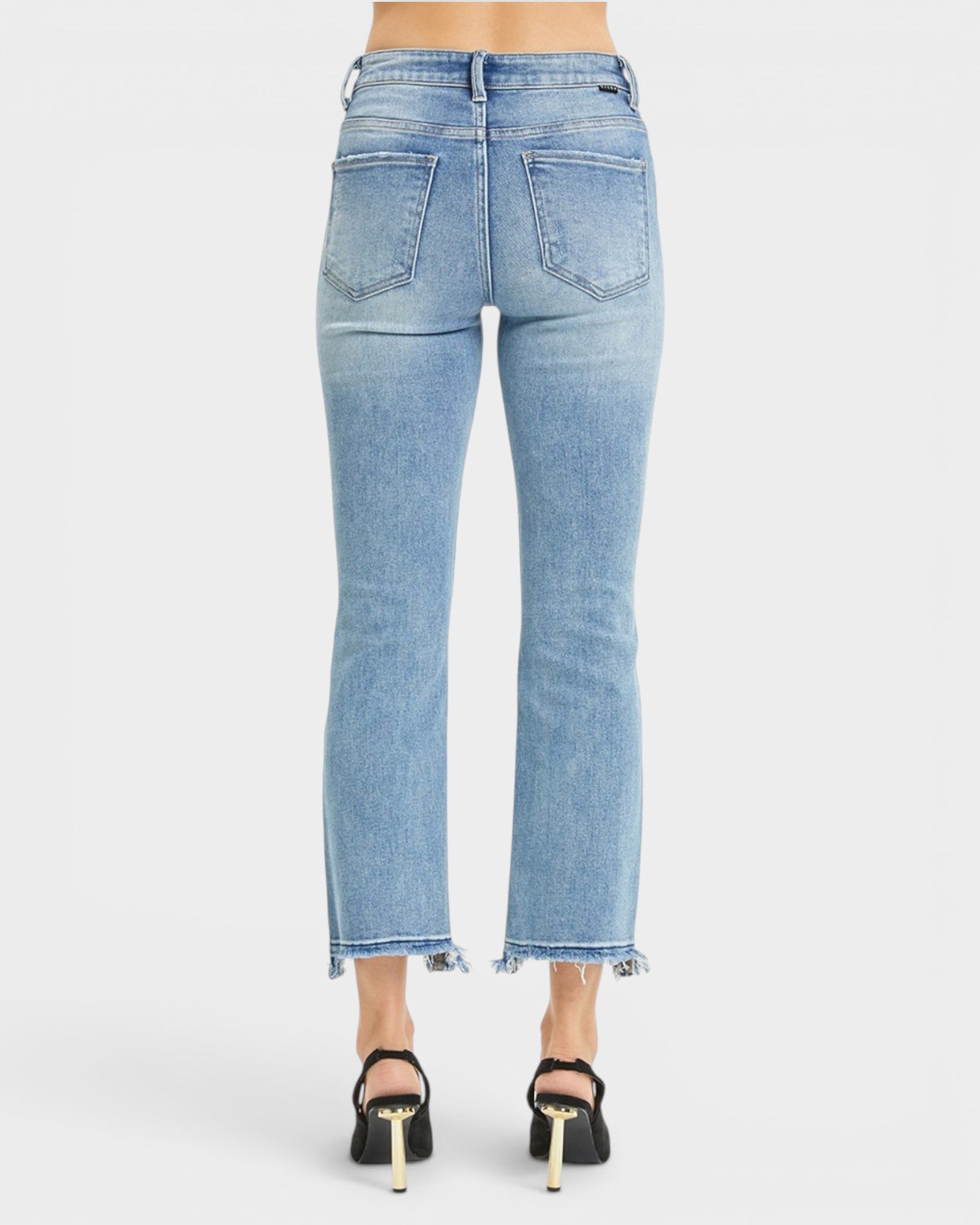 Back view of a model wearing light blue cropped flare jeans with a frayed hem and black high heels against a light gray studio background.