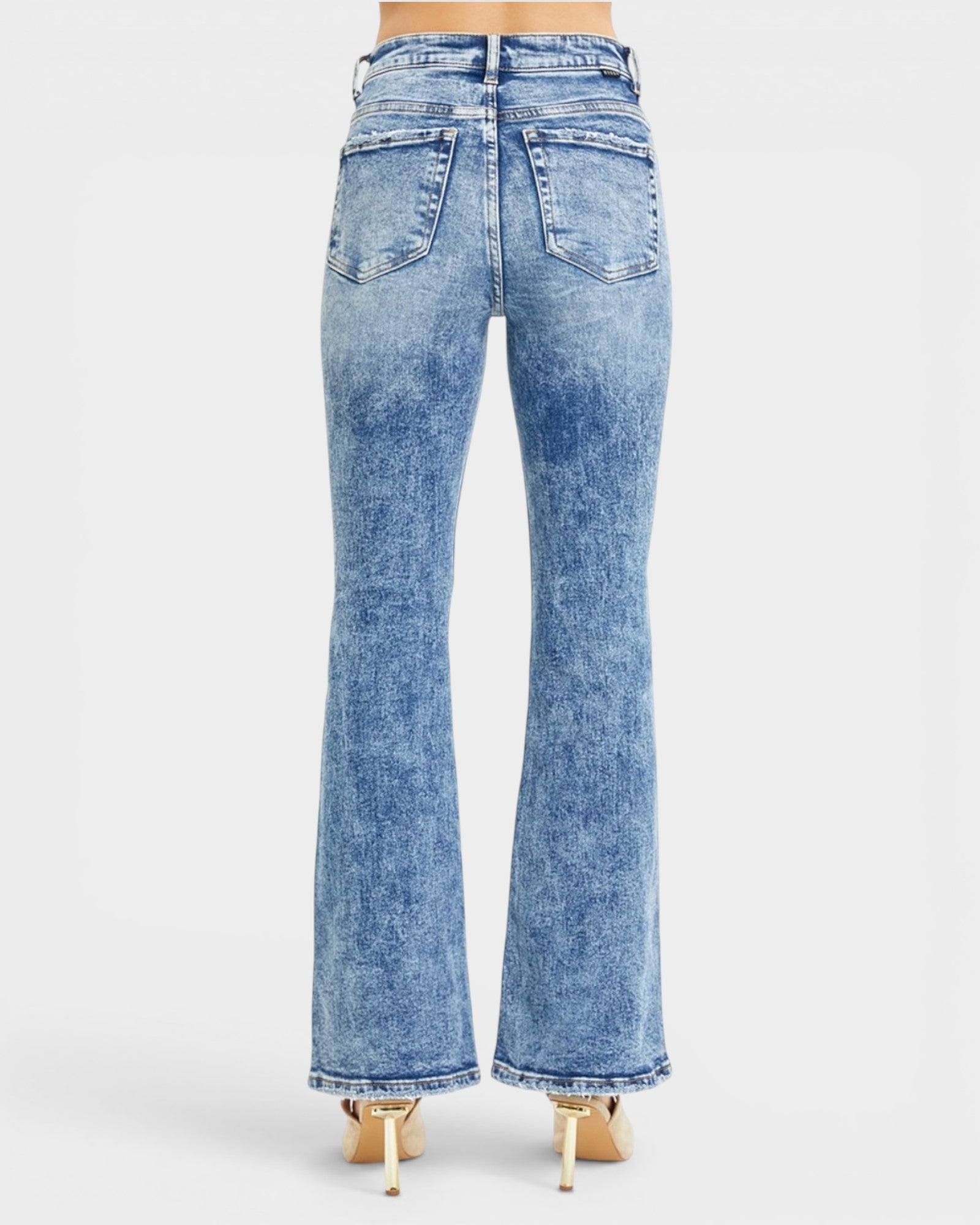 Rear view of a person wearing light blue high-waisted flare jeans with a mineral wash and gold high heels, shown against a light gray studio background.