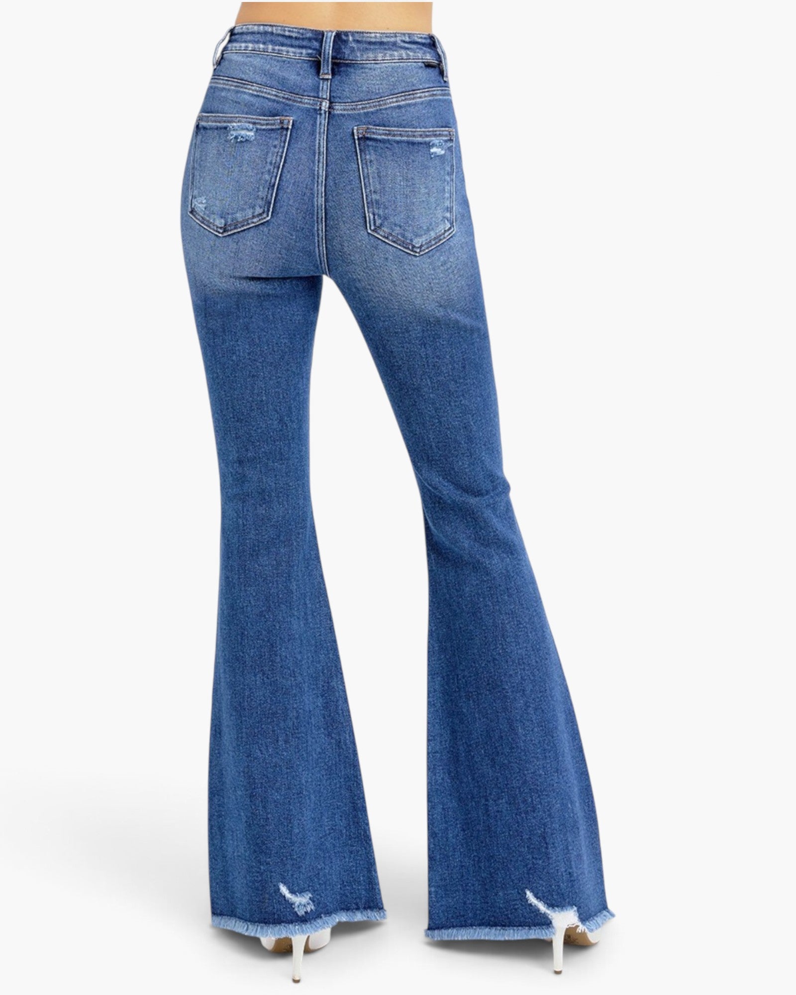 Rear view of a person wearing blue high-waisted flare jeans with distressed details and frayed hems, paired with white heels, against a light gray studio background.