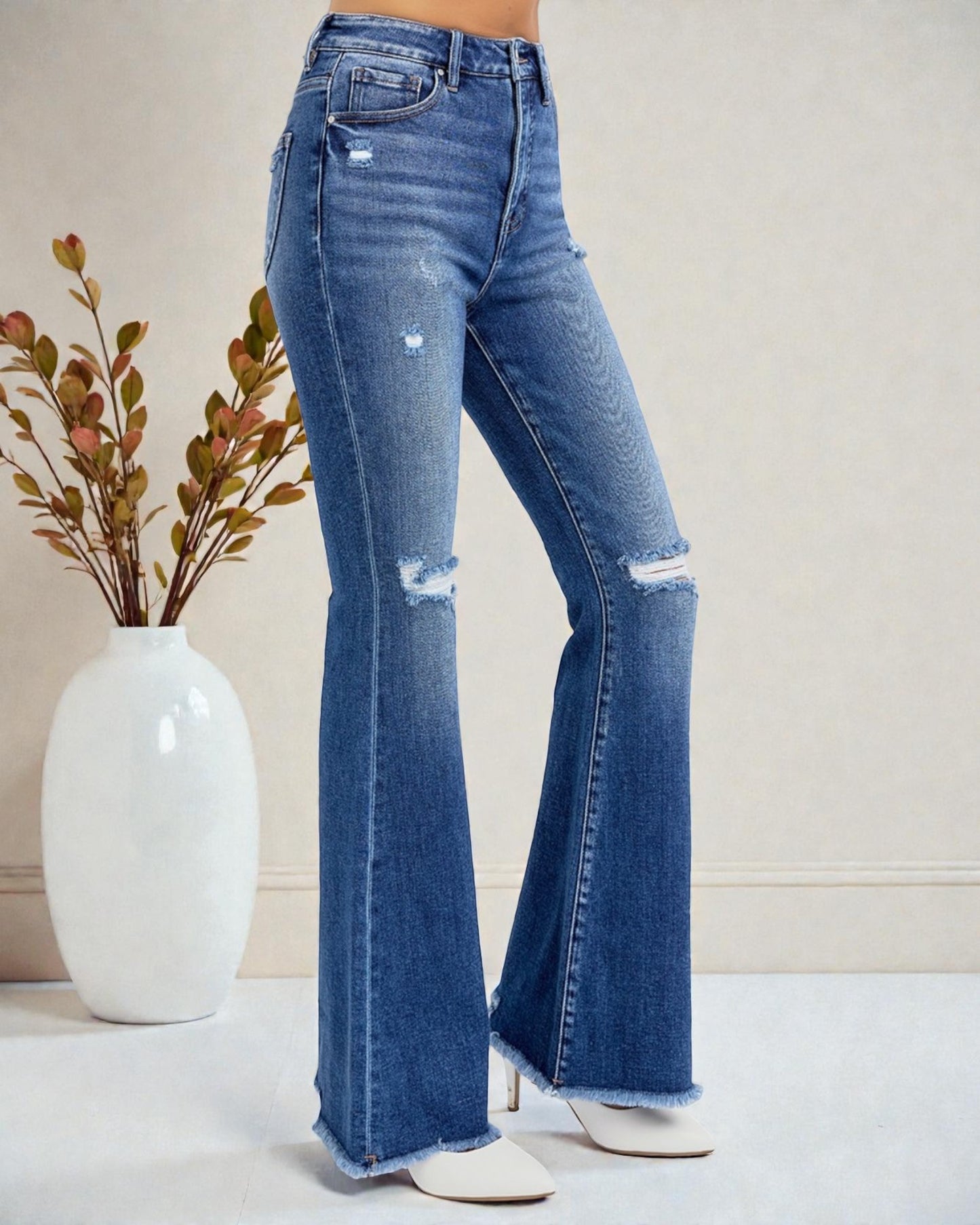 Blue distressed flared jeans worn by a person on a neutral background with a vase of flowers.