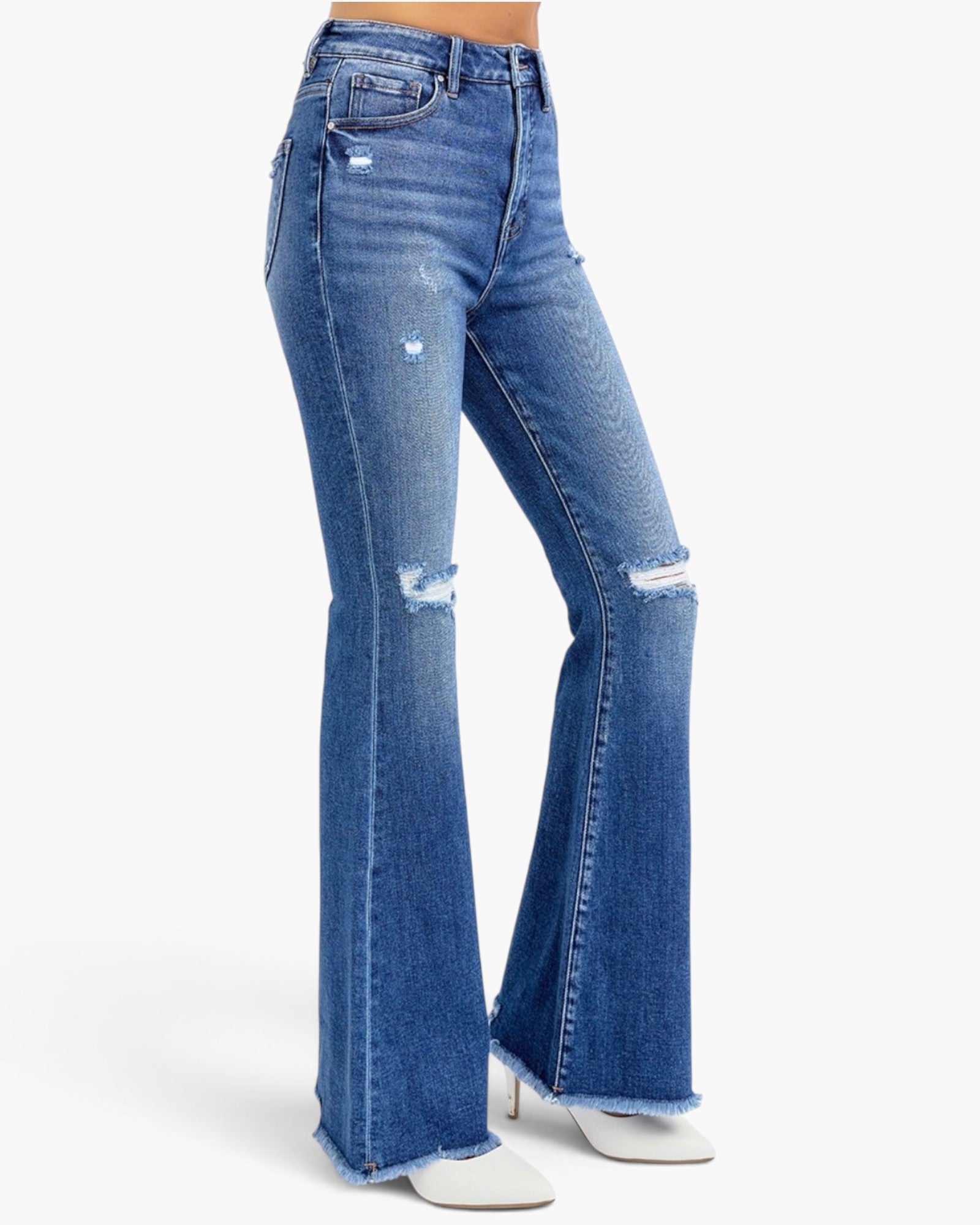 A model wearing a pair of medium-wash blue denim jeans with a high-rise, flared silhouette, featuring distressed details and rips on the thighs and knees, paired with white pointed-toe heeled boots against a plain white studio background.