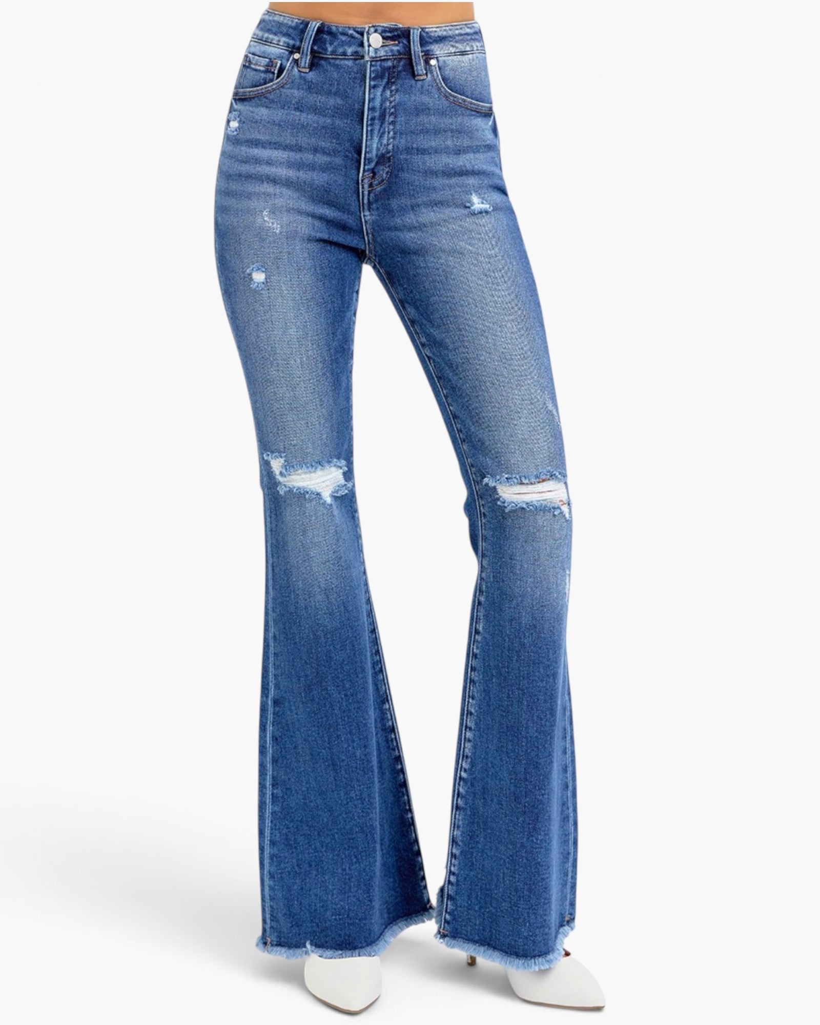 A pair of high-waisted, medium wash blue denim jeans with distressed details and a flared leg, shown on a model from the waist down wearing white pointed-toe heels against a light gray background.