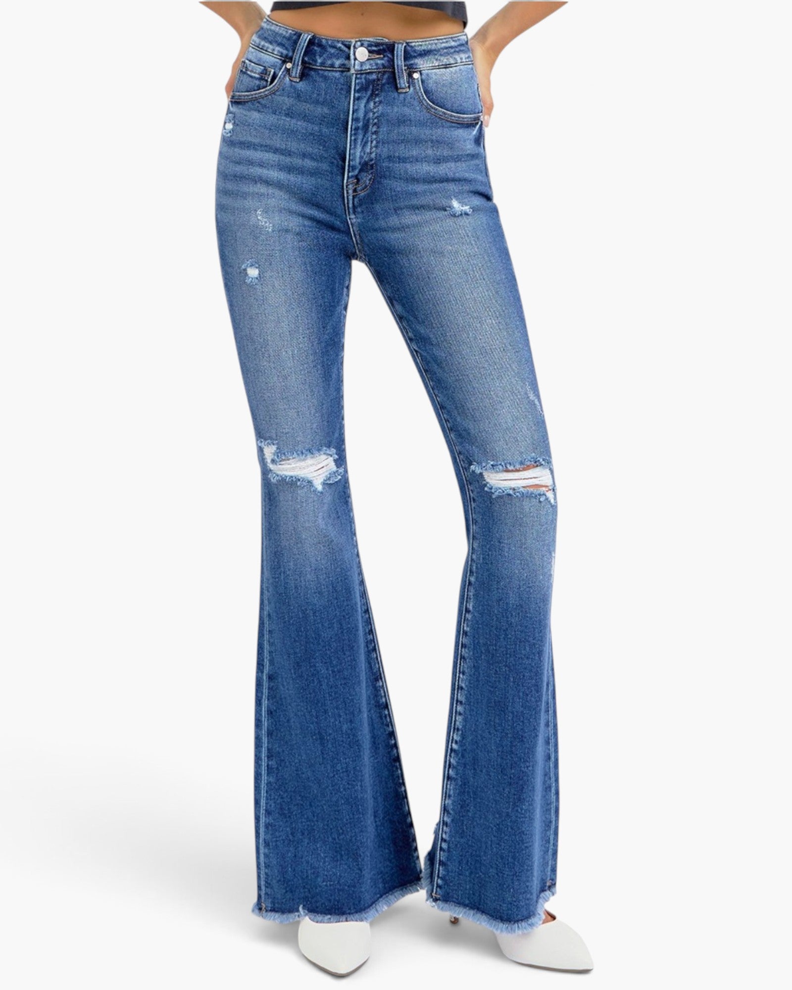 A pair of high-waisted, medium wash blue denim flare jeans with distressed details and frayed hems, shown on a model from the waist down wearing white pointed-toe heels against a light gray background.