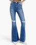 A pair of high-waisted, medium wash blue denim flare jeans with distressed details and frayed hems, shown on a model from the waist down wearing white pointed-toe heels against a light gray background.