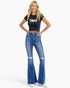 A woman modeling a black graphic crop top and high-waisted, distressed blue flare jeans with white sneakers, set against a plain white studio background.