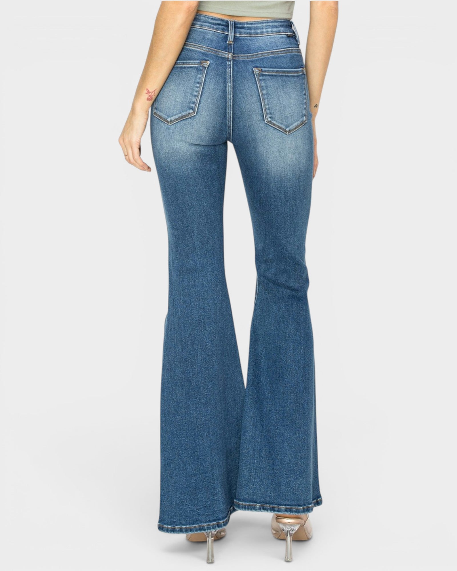 Rear view of a model wearing blue denim flare jeans with a high waist and wide flared legs, paired with metallic high heels against a light gray background.