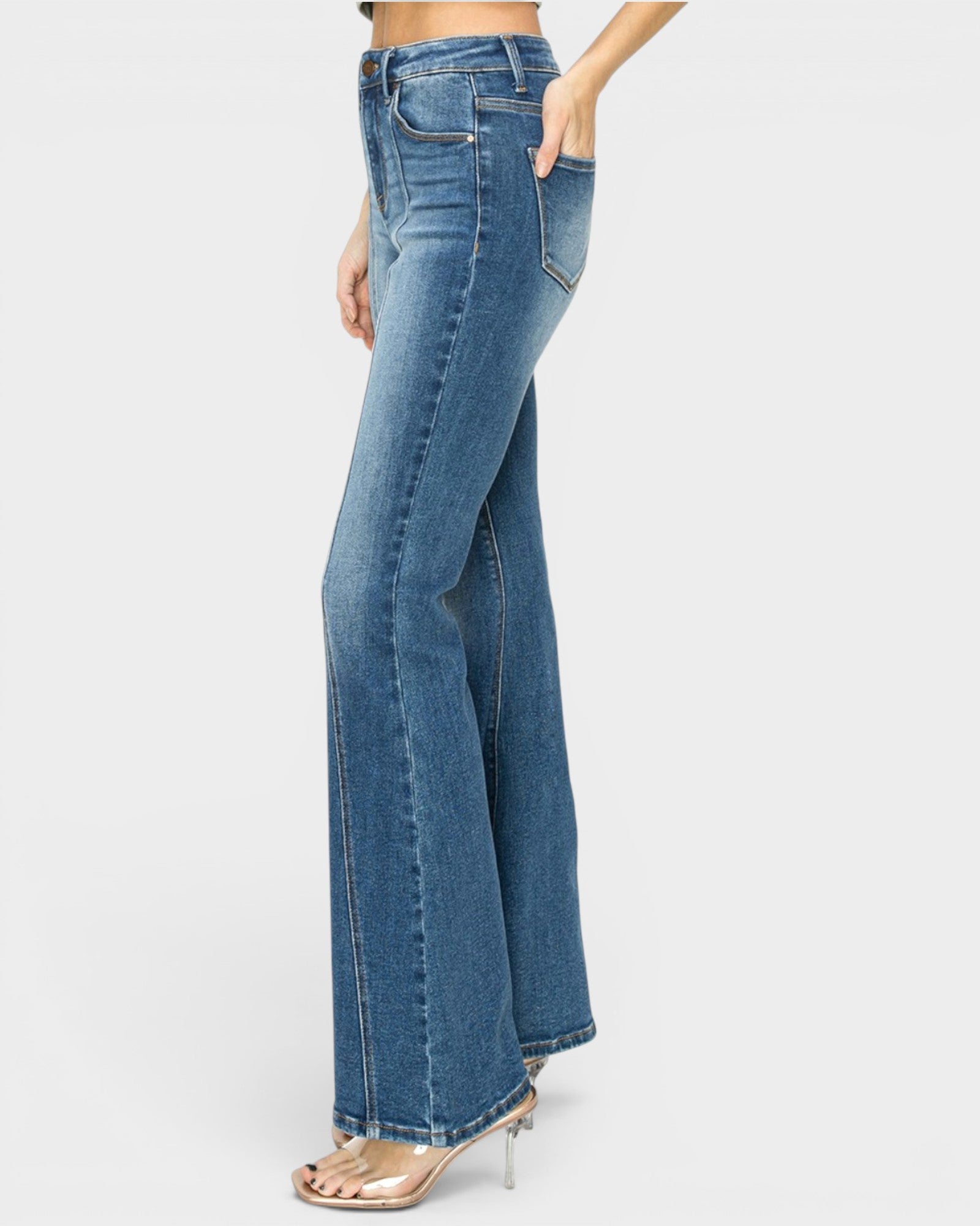 A side view of a model wearing high-waisted, wide-leg blue denim jeans with a side pocket, paired with clear high-heeled sandals, against a light gray background.