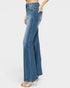 A side view of a model wearing high-waisted, wide-leg blue denim jeans with a side pocket, paired with clear high-heeled sandals, against a light gray background.