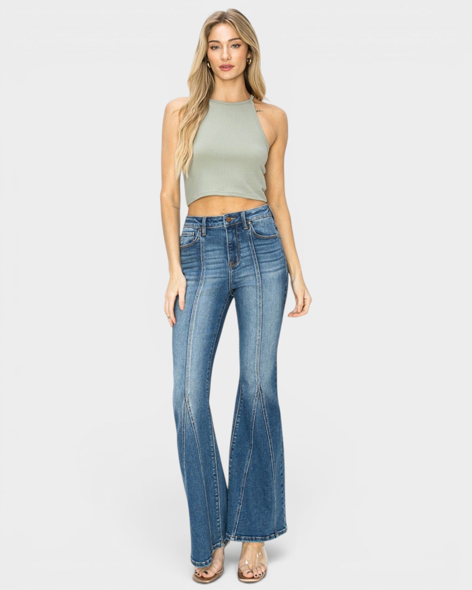 A woman modeling a sleeveless sage green crop top and blue high-waisted flare jeans against a light gray studio background.