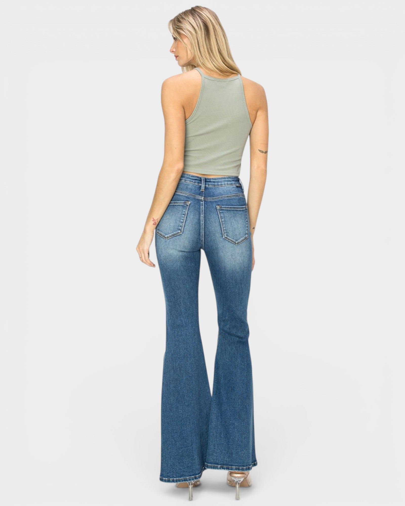 A woman shown from the back wearing blue high-waisted flare jeans and a sage green sleeveless crop top, standing against a light gray studio background.