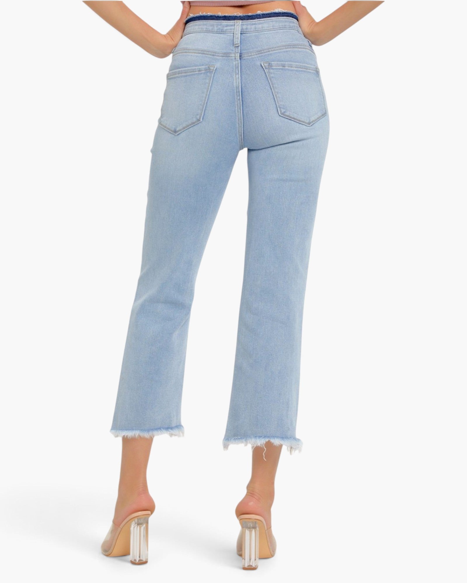 Back view of a model wearing light-wash blue cropped jeans with a frayed hem and a dark blue waistband, paired with clear-strap high-heeled sandals against a light gray studio background.
