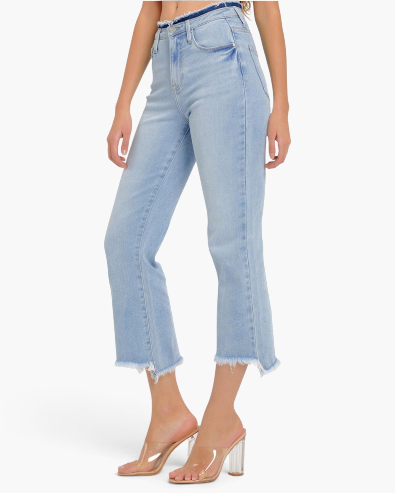 A model wearing light blue cropped denim jeans with a raw frayed hem and clear high-heel mules, shown against a plain white studio background.