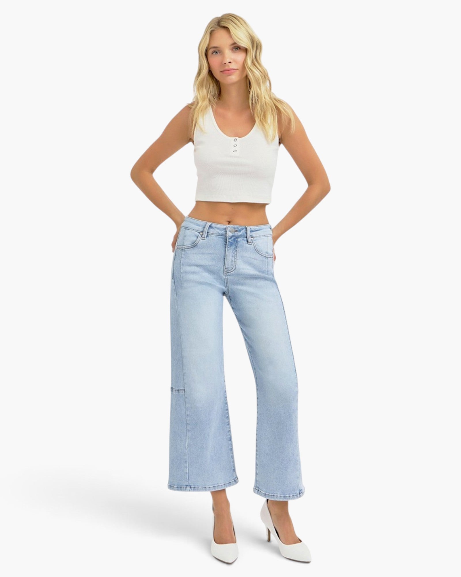 A blonde model wearing a white ribbed crop top, light-wash wide-leg cropped jeans, and white pointed-toe heels, standing against a light gray studio background.