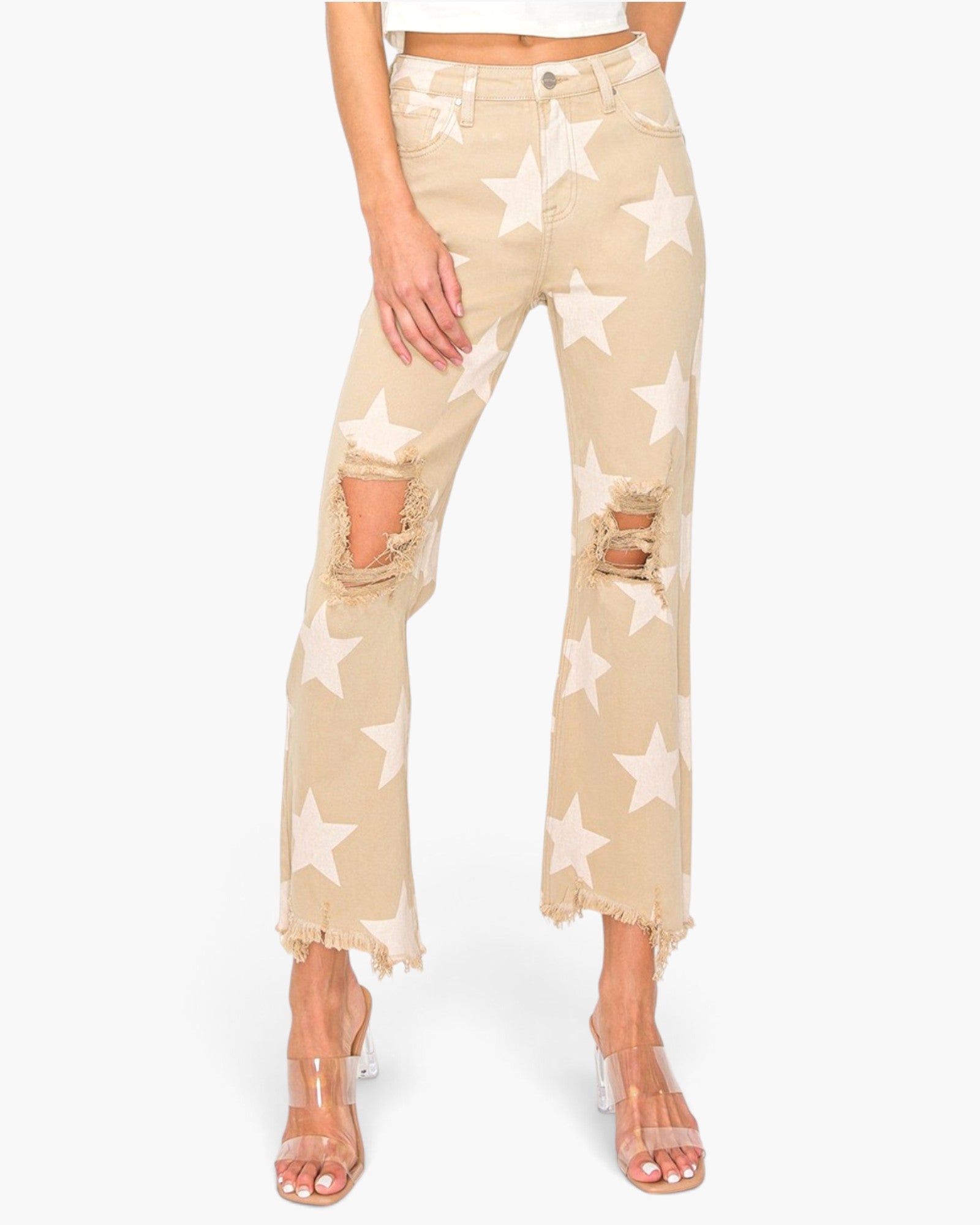 A model wearing beige cropped jeans with a white star print, distressed rips at the knees, and frayed hems, paired with clear high-heeled sandals against a light gray background.