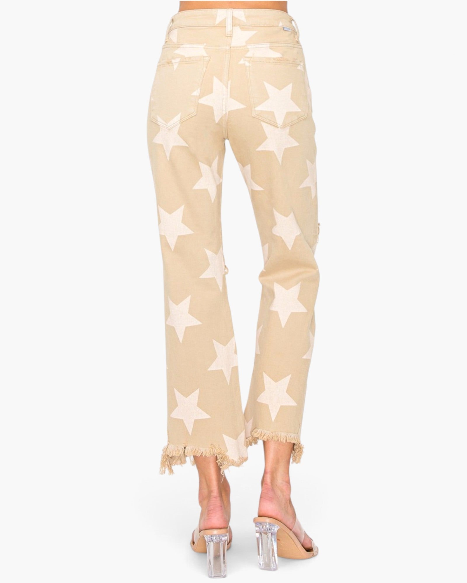 Back view of a model wearing beige cropped jeans with a white star pattern and frayed hems, paired with clear high-heeled sandals.