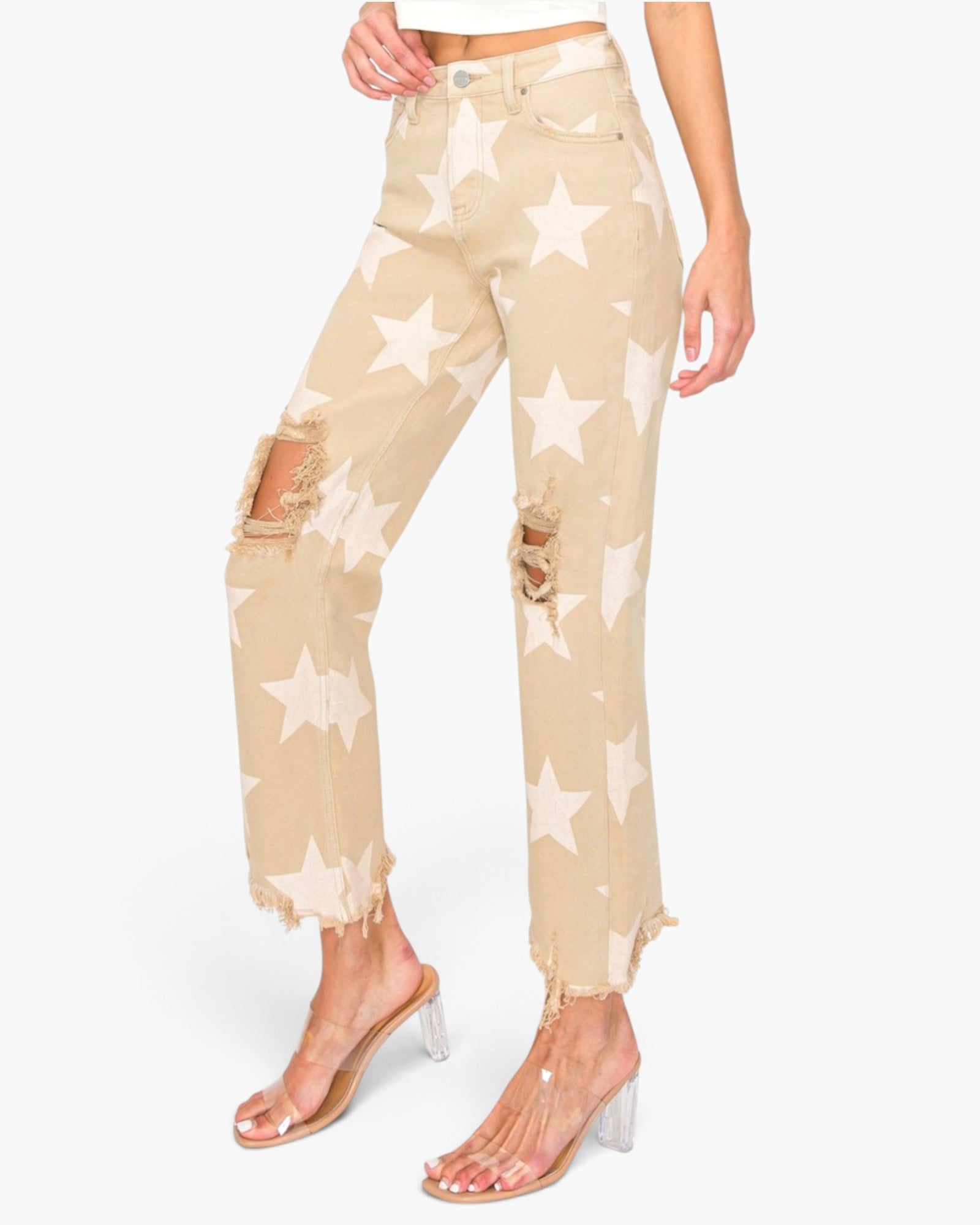 A model wears beige cropped jeans with a white star pattern and distressed rips on the knees, paired with clear high-heeled sandals, against a plain white background.