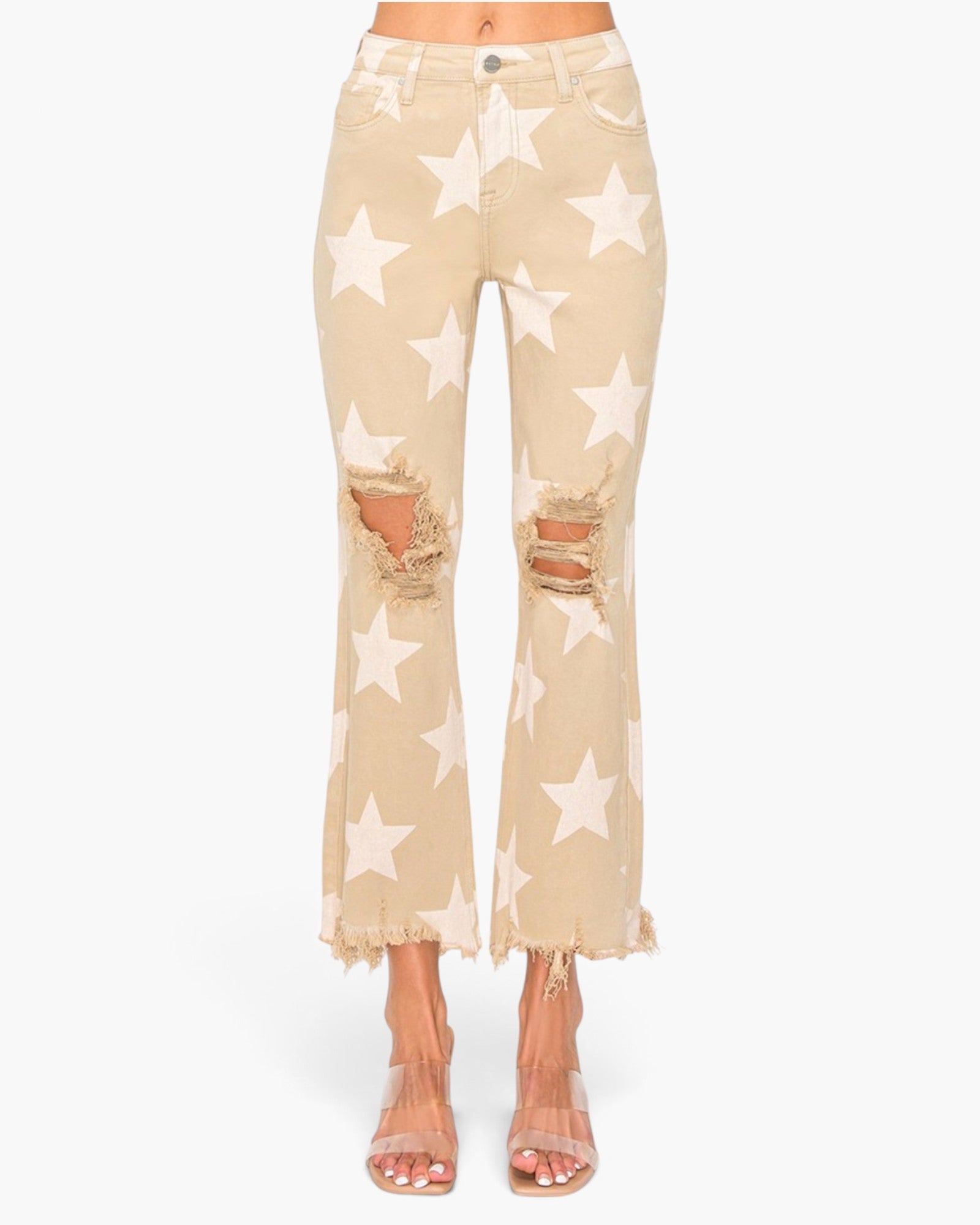 A pair of beige cropped flare jeans with a white star pattern, featuring distressed rips at the knees and a frayed hem, shown on a model wearing clear high-heeled sandals.