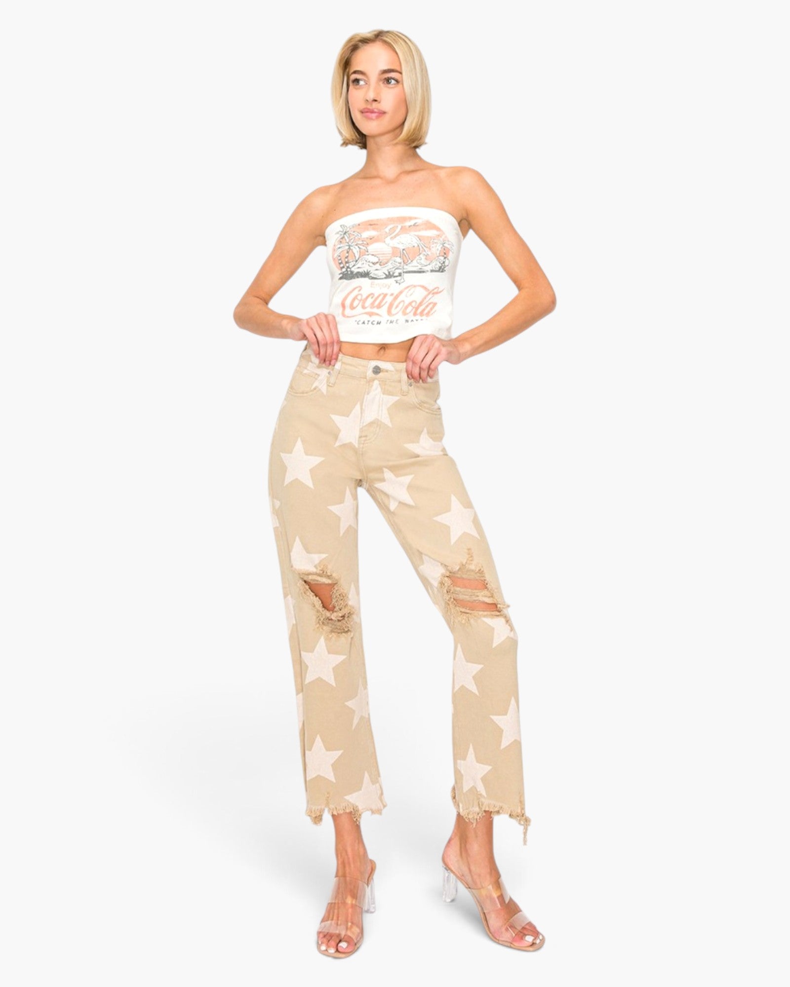 A woman modeling beige distressed jeans with a white star print, paired with a white strapless crop top and clear high-heeled sandals, set against a plain white background.