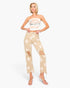 A woman modeling beige distressed jeans with a white star print, paired with a white strapless crop top and clear high-heeled sandals, set against a plain white background.