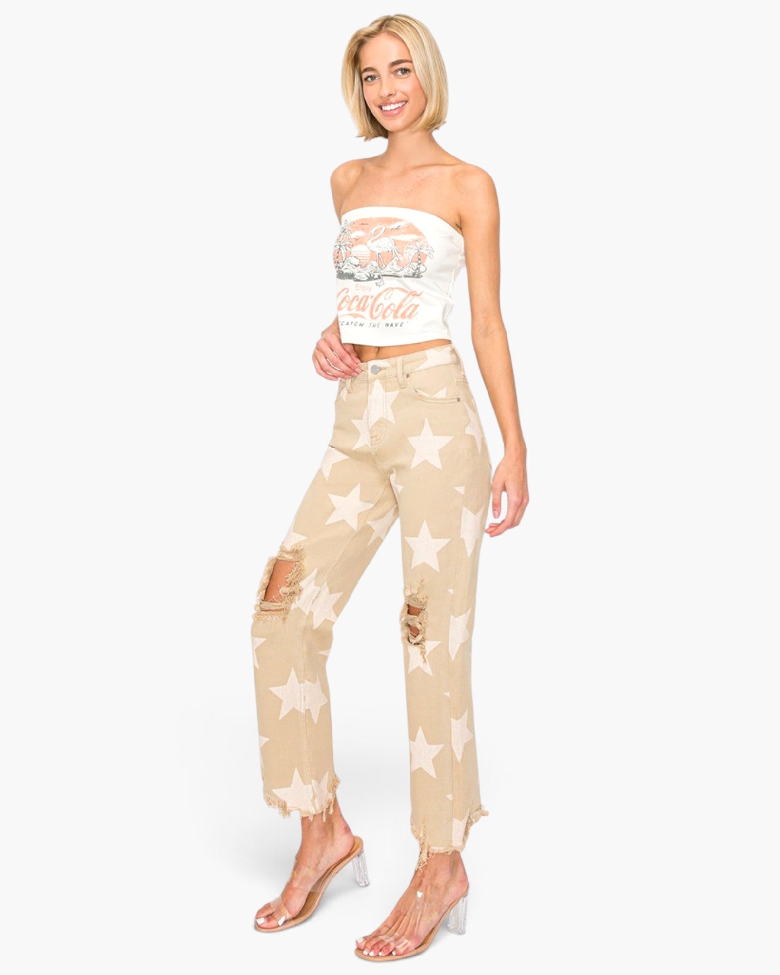 A woman modeling beige distressed jeans with a white star print, paired with a white strapless crop top and clear high-heeled sandals, set against a plain white background.