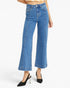 A model wearing high-waisted, wide-leg blue denim jeans with a cropped hem, styled with black pointed-toe slingback heels against a clean white studio background.