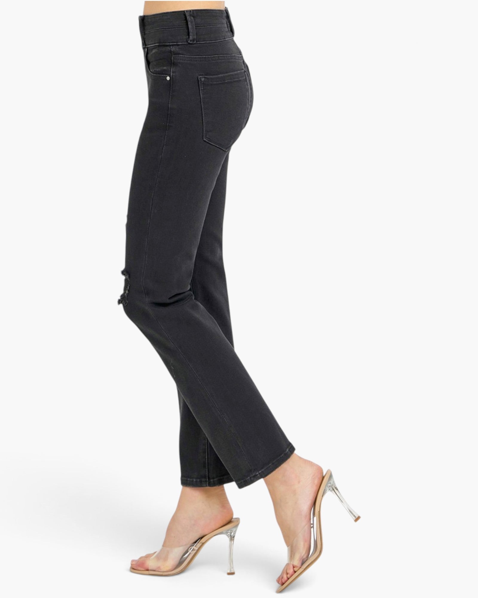 A side profile view of a model wearing black cropped jeans and clear strap high-heeled sandals against a plain white background.