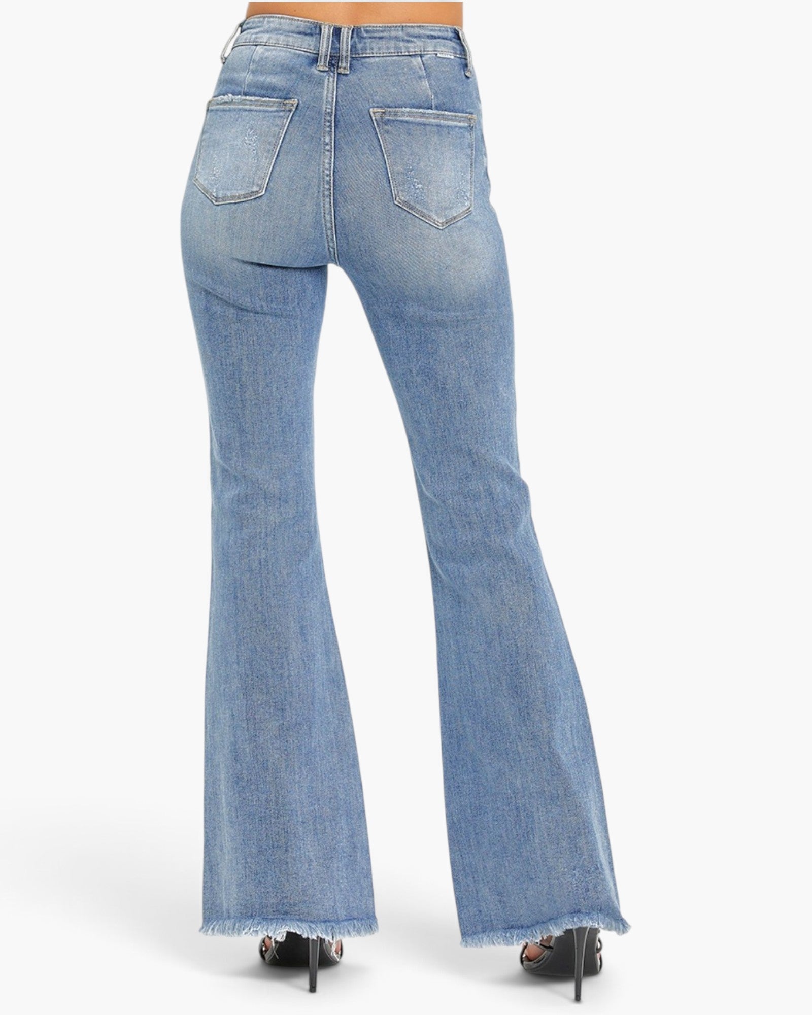 Rear view of a person wearing light-wash blue flared denim jeans with a frayed hem, paired with black high heels, against a plain light gray studio background.