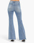 Rear view of a person wearing light-wash blue flared denim jeans with a frayed hem, paired with black high heels, against a plain light gray studio background.