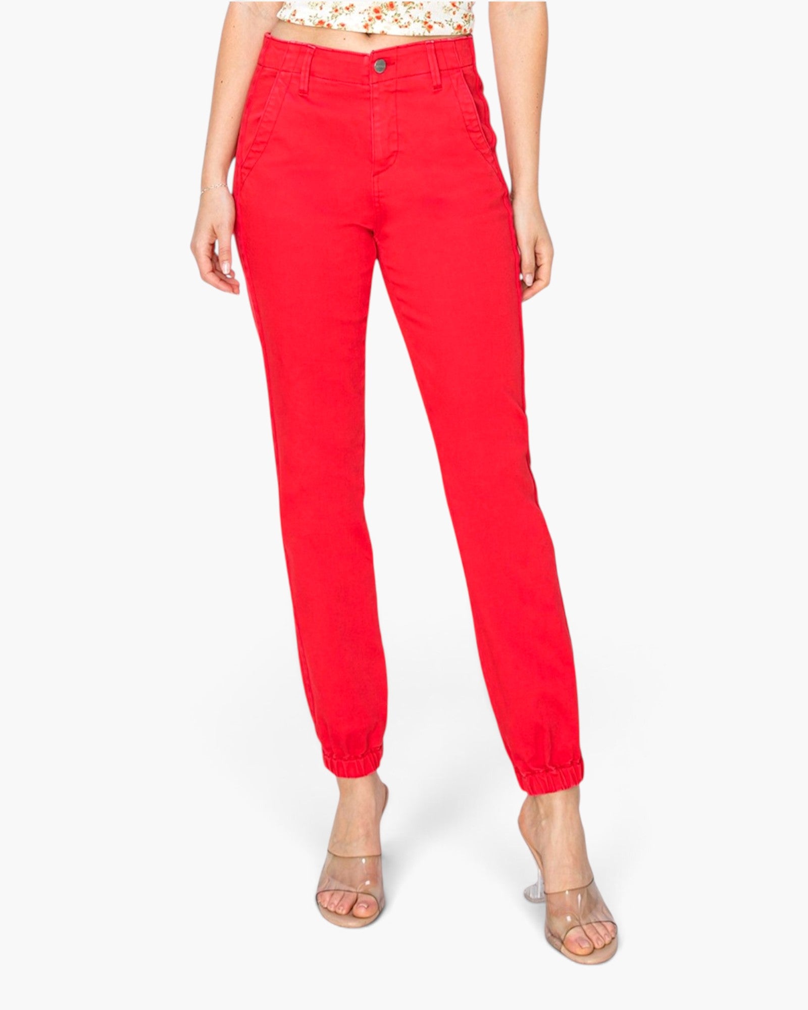 A model wearing bright red ankle-length pants with a gathered elastic hem, paired with clear heeled sandals and a floral top, shown against a plain white background.