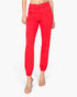 A model wearing bright red jogger-style pants with elastic cuffs, paired with clear heeled sandals, shown against a plain light gray studio background.