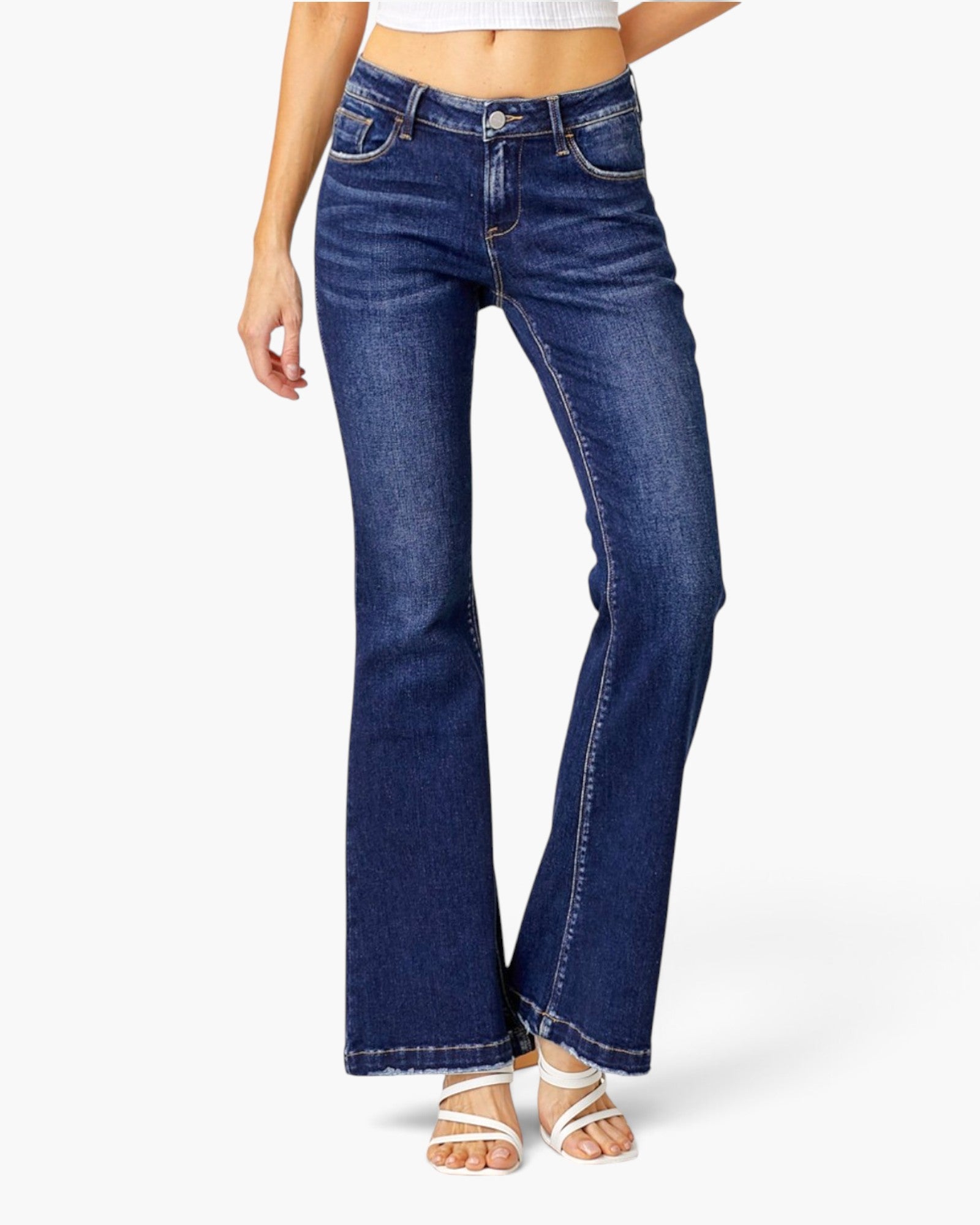 A model wearing dark blue low-rise flare jeans with a button fly and contrast stitching, paired with white strappy sandals against a light gray background.