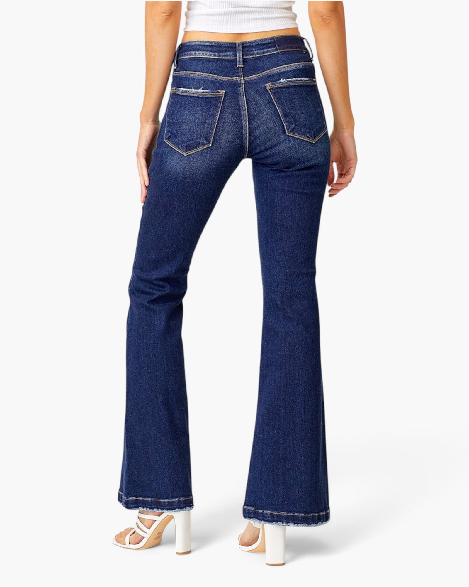 Rear view of a model wearing dark blue flare jeans with a white crop top and white high-heeled sandals against a light gray studio background.