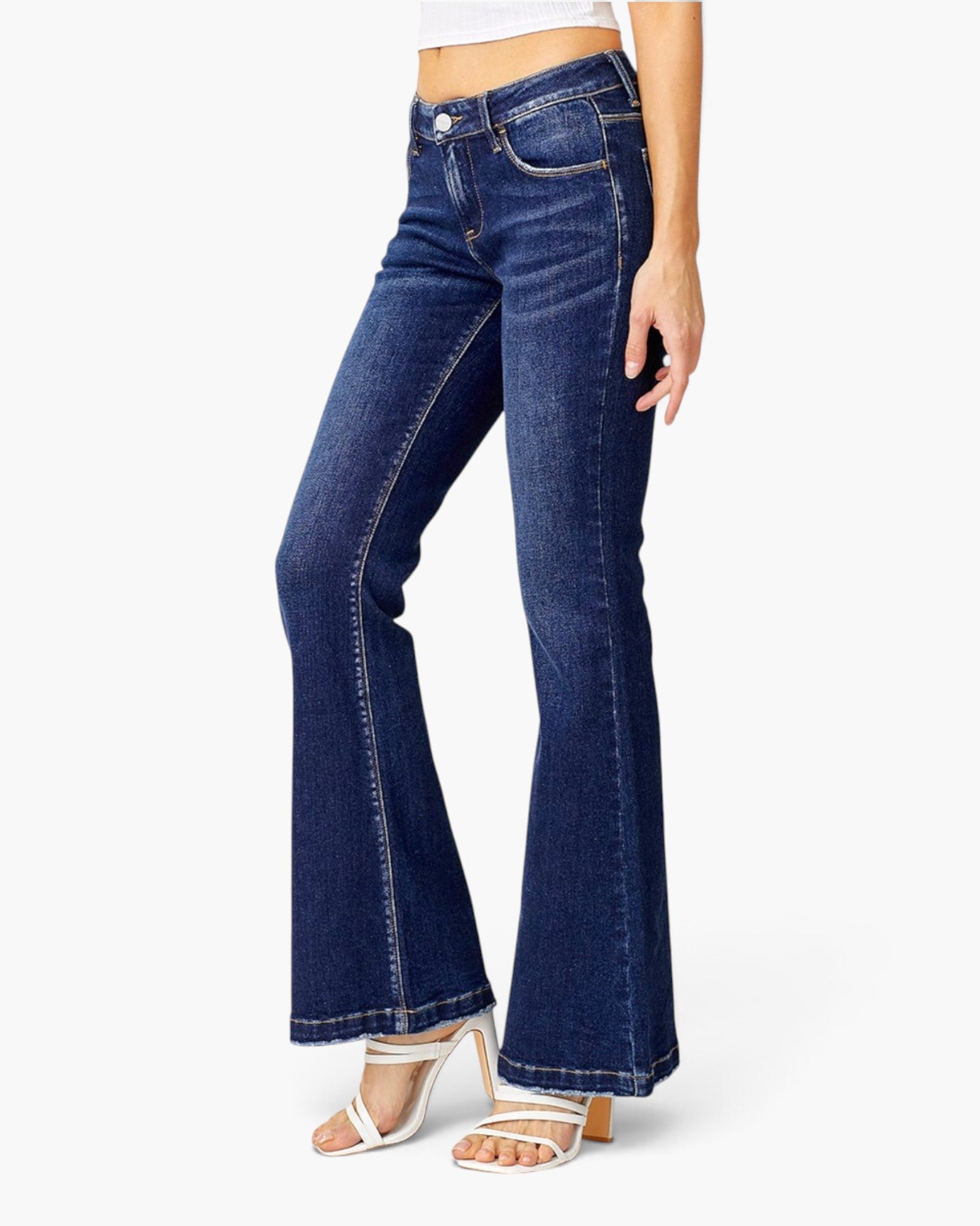 A model wearing dark blue flare jeans with white strappy high-heeled sandals, shown from the waist down on a light gray background.
