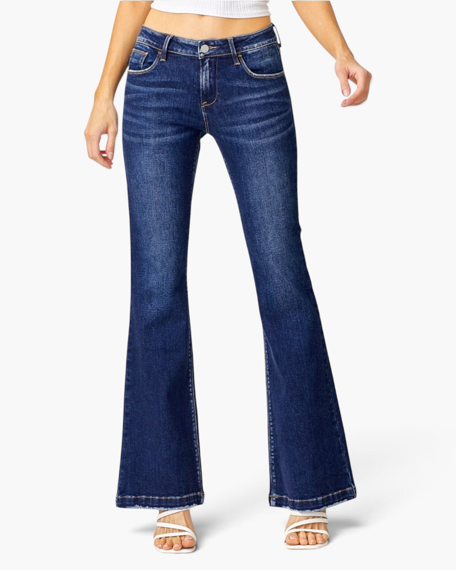 A model wearing a pair of dark blue low-rise flare jeans with a subtle whiskering effect on the thighs, paired with a white crop top and white heeled sandals, against a light gray studio background.