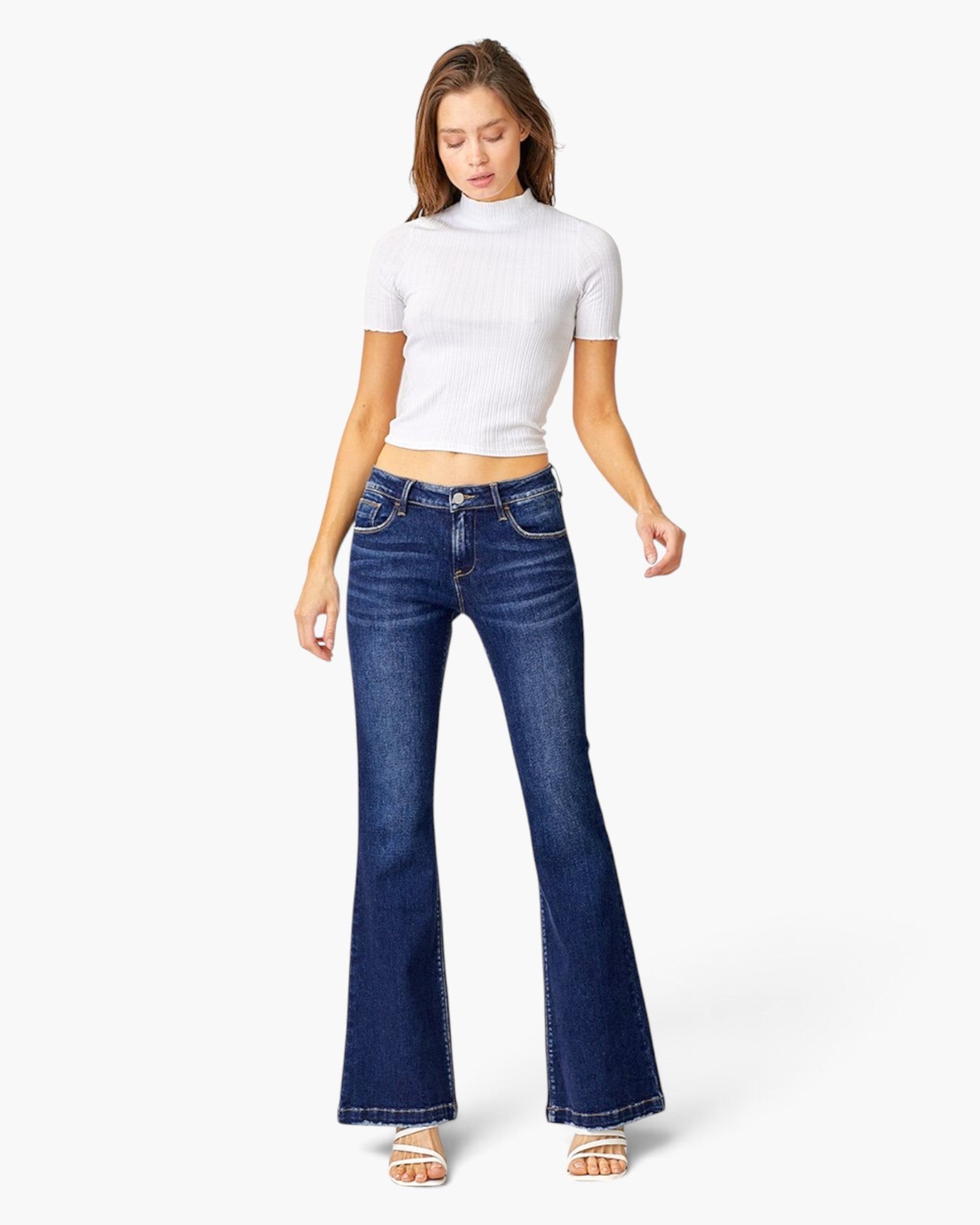 A woman modeling a pair of dark blue low-waisted flare jeans with a white short-sleeve crop top, standing against a light gray studio background.
