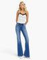 A blonde model wearing a white camisole top with black lace trim and blue high-waisted flare jeans, standing against a plain white studio background.