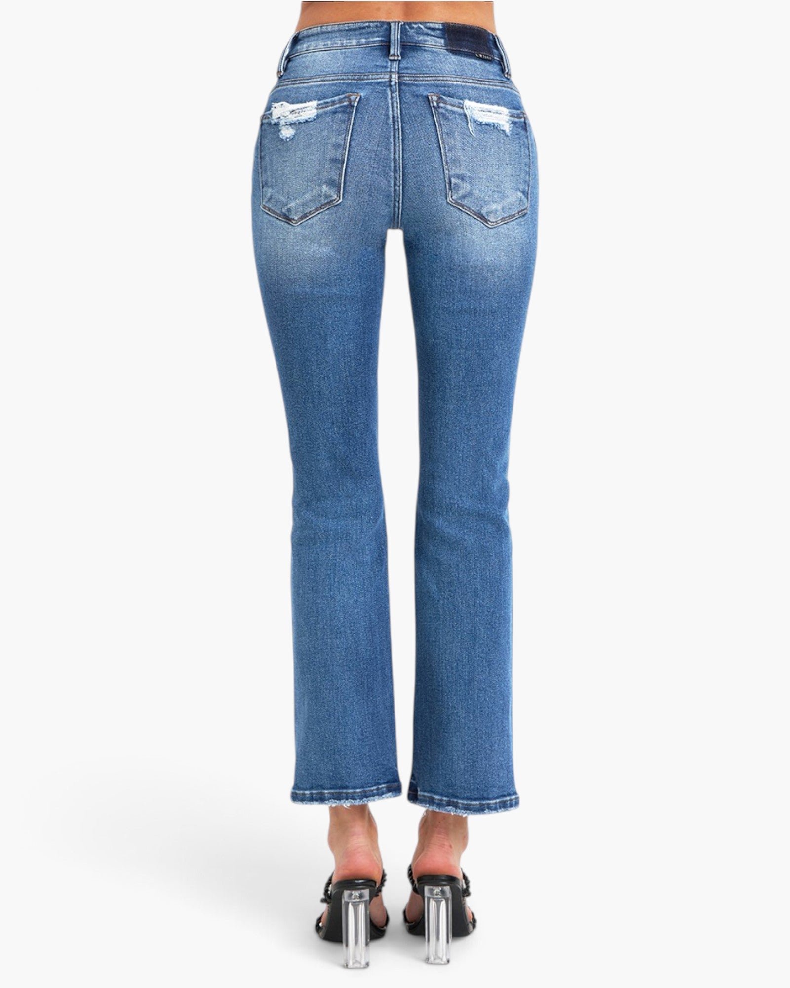 Rear view of a model wearing blue denim flare jeans with distressed details on the back pockets and frayed hems, paired with black high-heeled sandals against a plain white background.