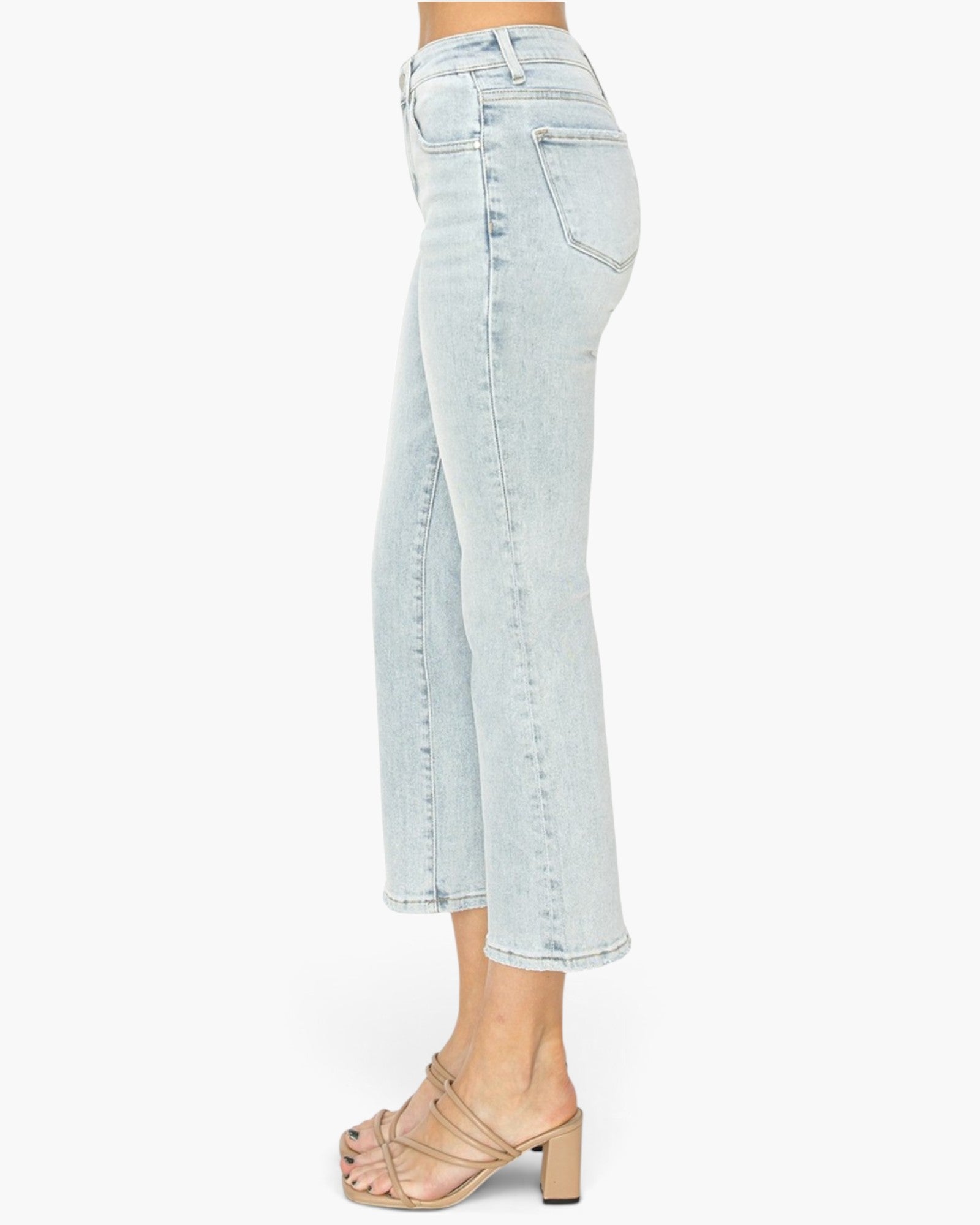 Side view of a person wearing light-wash cropped denim jeans and beige strappy heeled sandals against a plain white background.