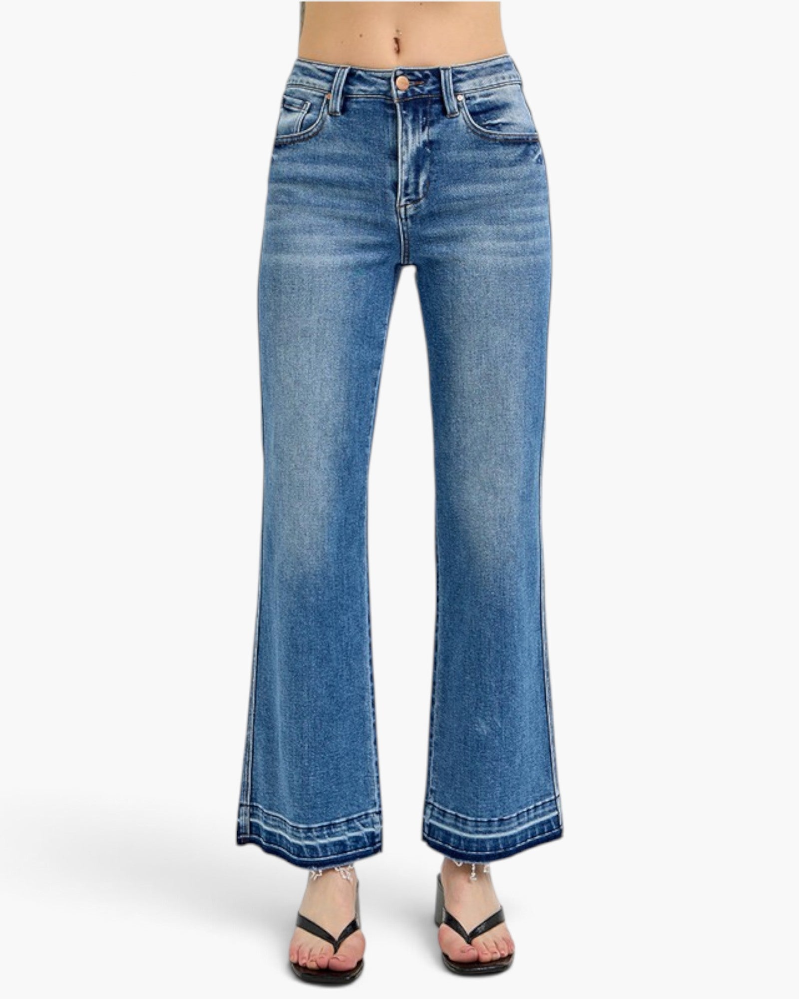 A person wearing medium-wash blue denim flare jeans with a straight rise and wide legs, paired with black flip-flop sandals, shown against a plain light gray studio background.