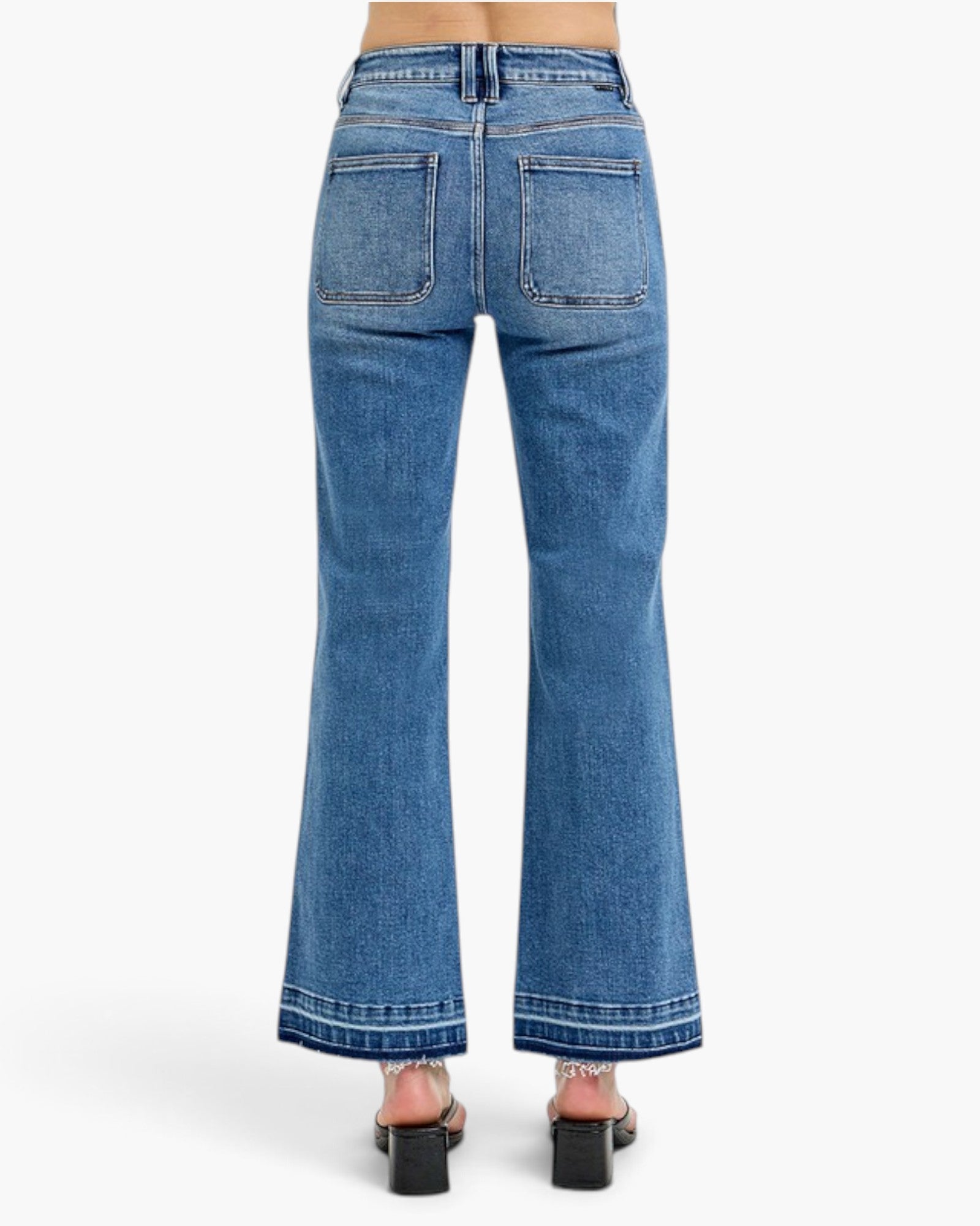Rear view of a person wearing blue wide-leg denim jeans with a straight-to-flared leg and black block-heel sandals against a light gray studio background.