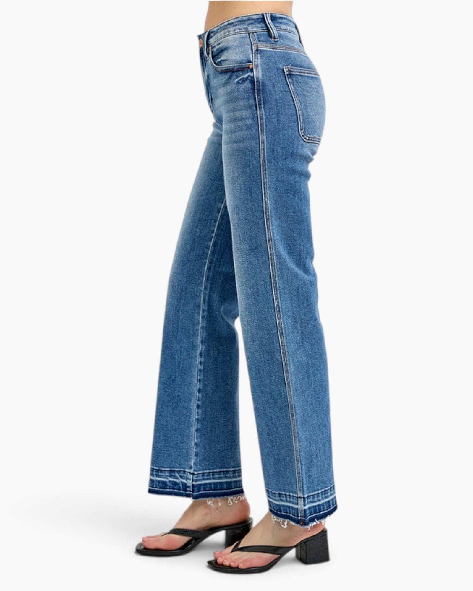 A person wearing blue wide-leg jeans with a straight-to-slightly-flared leg, shown from the waist down against a clean white studio background. The model is wearing black heeled sandals.