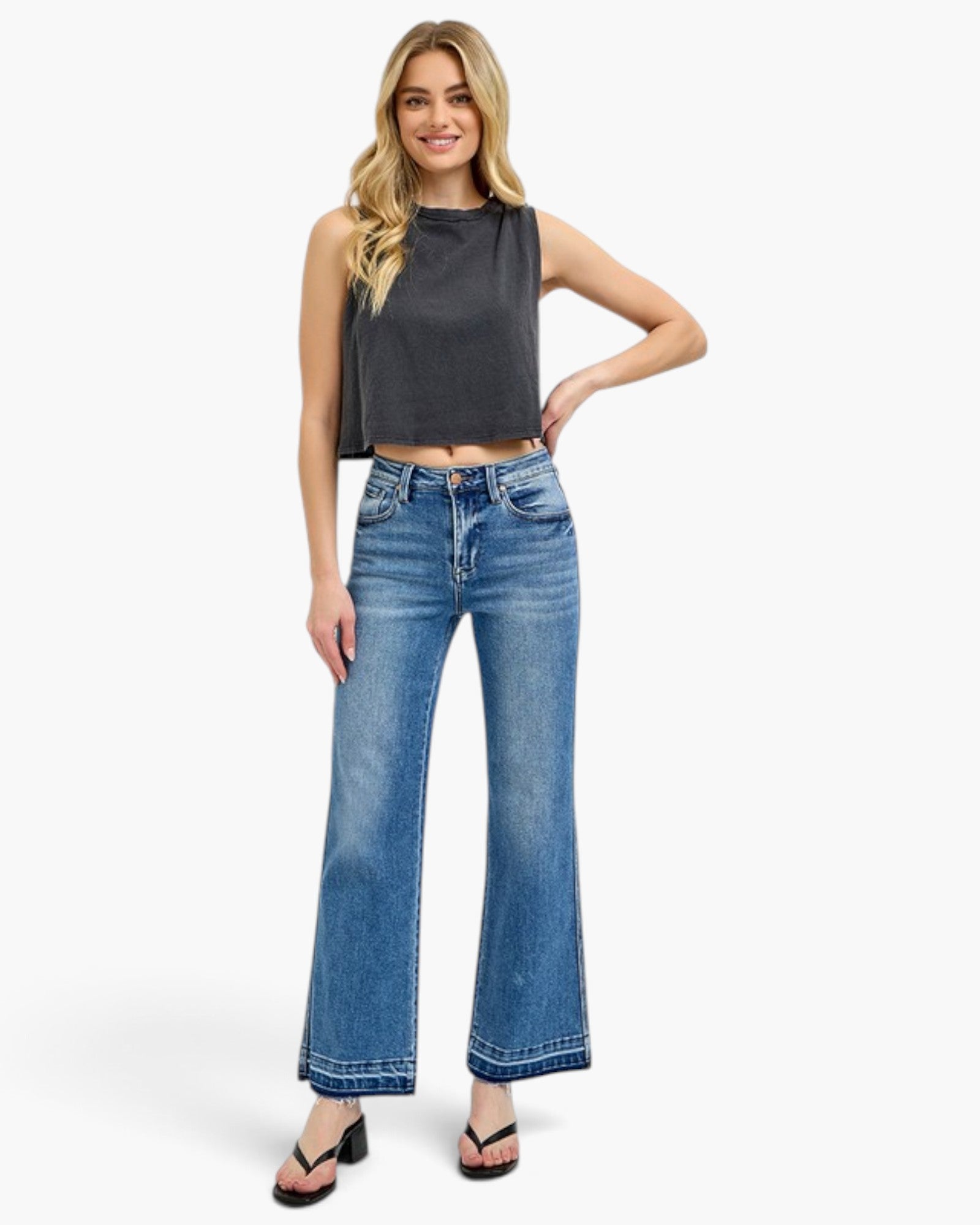 A woman modeling a dark charcoal gray sleeveless crop top and high-waisted, wide-leg blue denim jeans with a faded wash, paired with black sandals, against a plain white studio background.